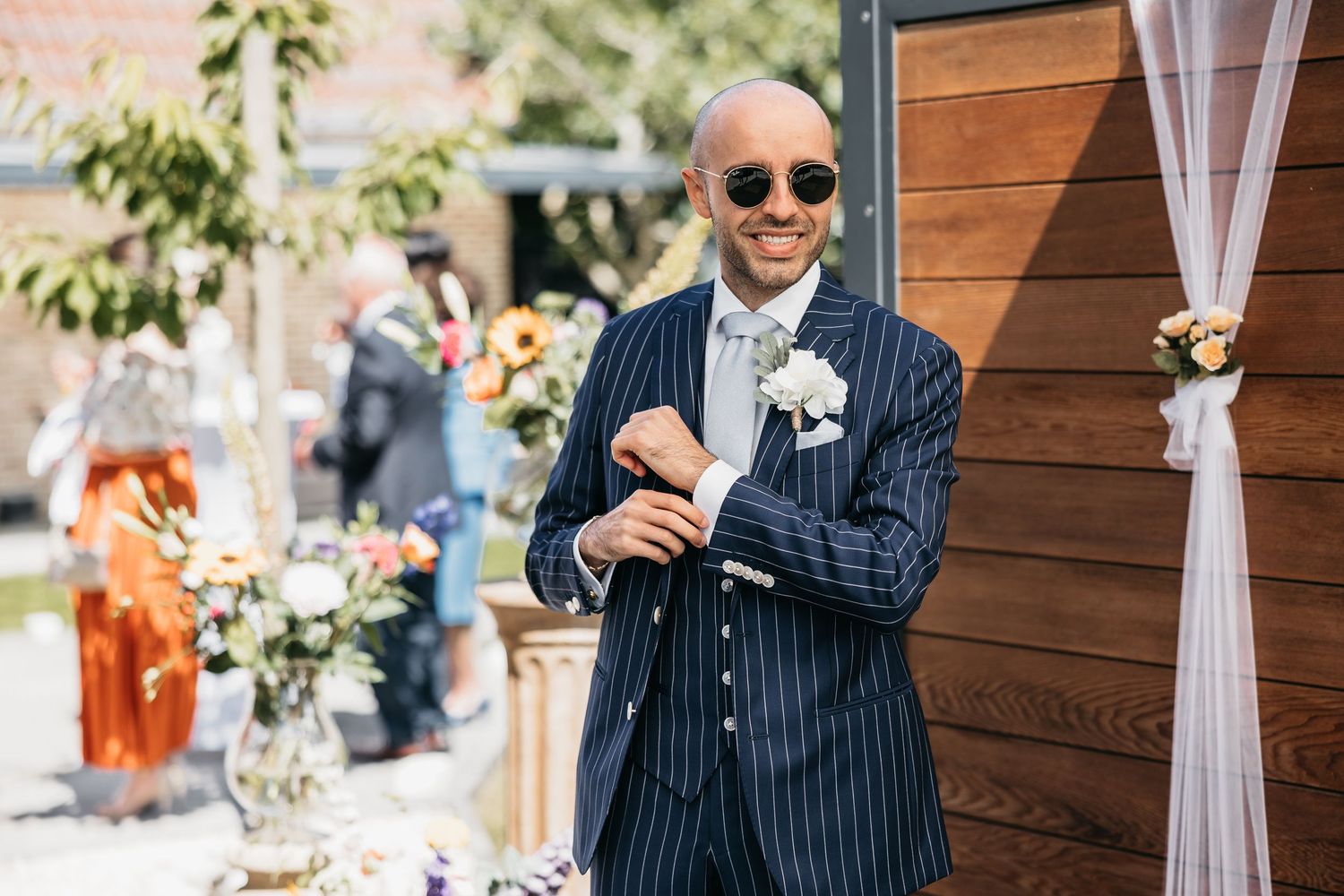 Stylish bald groom smiling in navy pinstripe suit with boutonniere and round sunglasses outdoors.