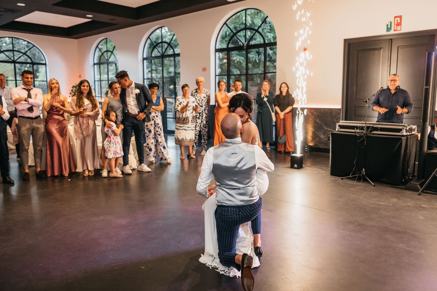 Wedding couple sharing first dance while guests including children watch in elegant venue with arched windows.