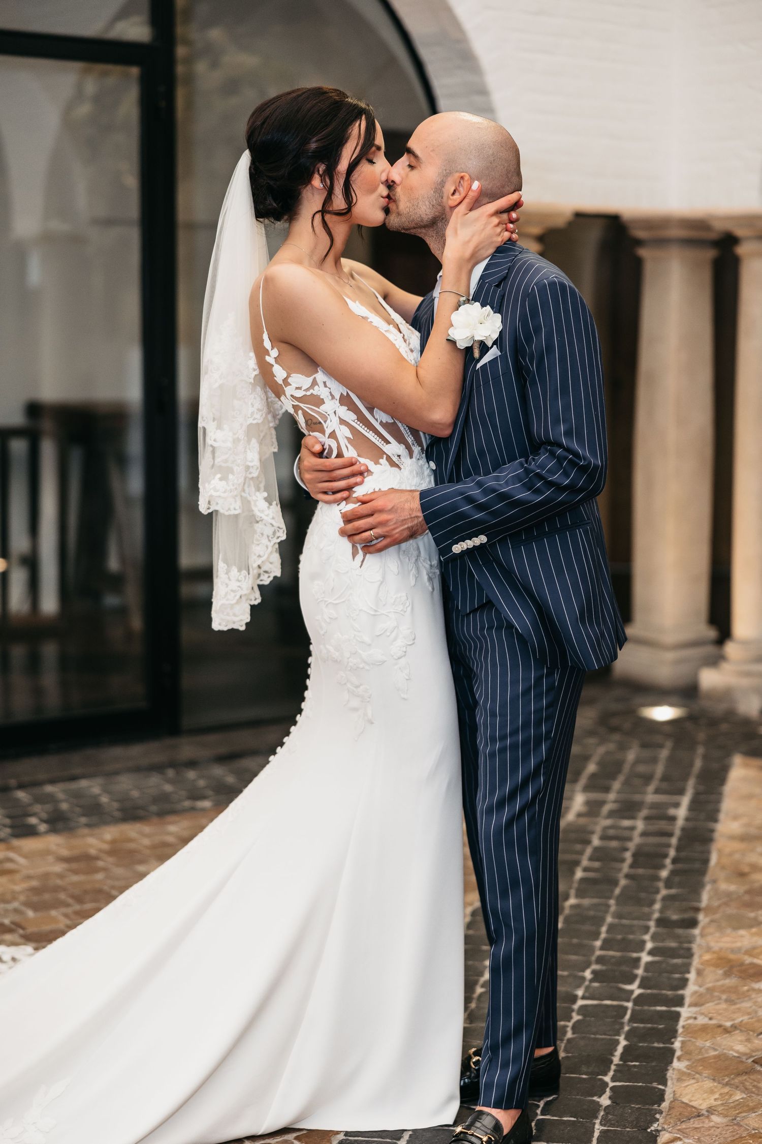 Bride in fitted lace wedding dress and groom in pinstripe suit share romantic kiss outdoors.