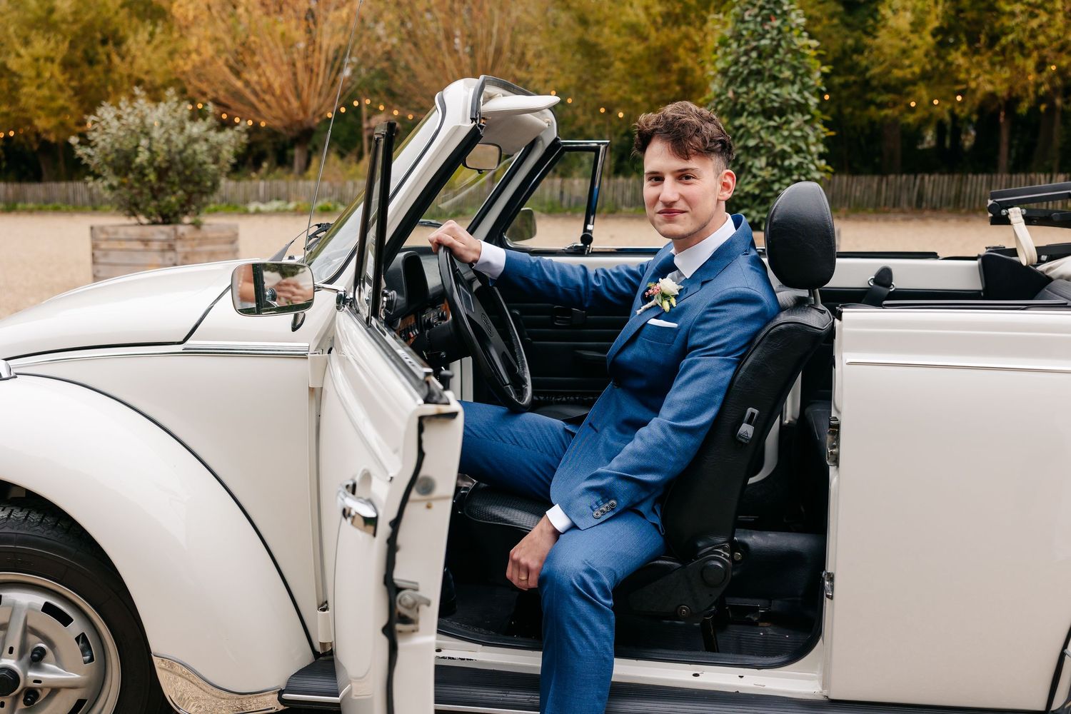 Dapper groom in blue suit posing beside open door of classic white convertible wedding car.