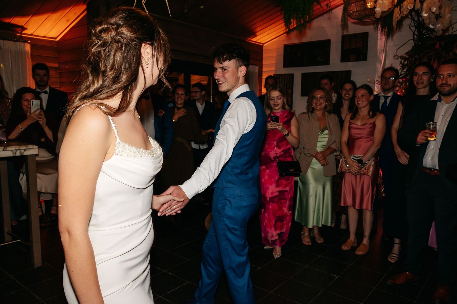 Bride and groom hold hands during first dance at wedding reception with smiling guests watching in background.