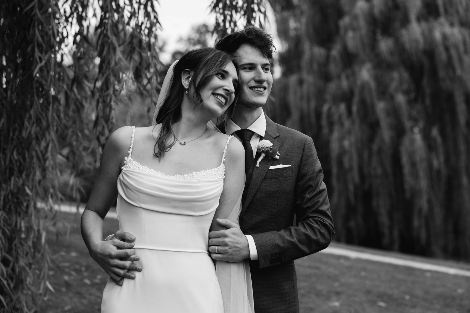 Black and white photo of laughing bride and groom embracing under weeping willow on wedding day.