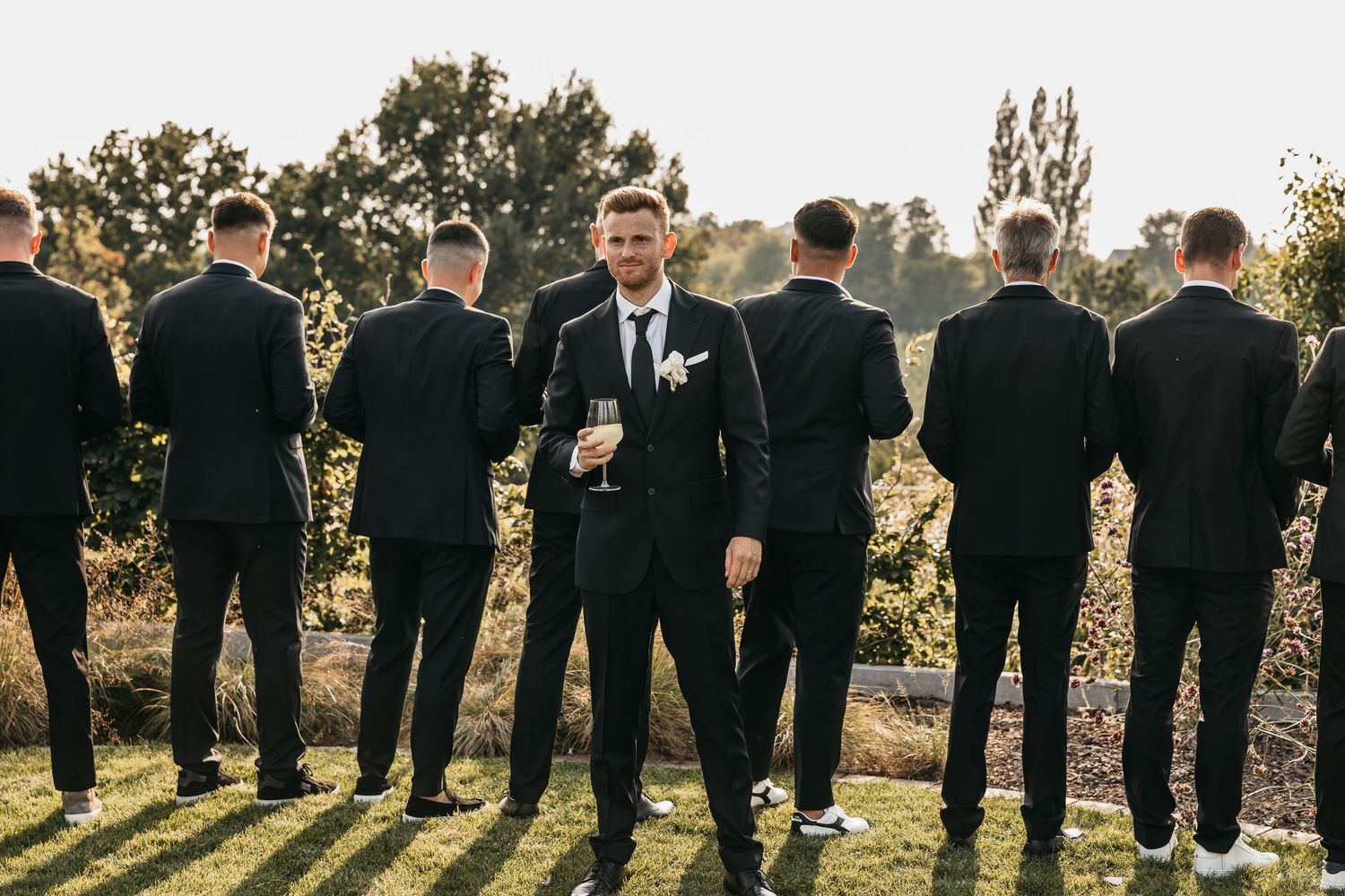 Groom holding champagne glass stands before groomsmen lined up on green lawn at wedding.