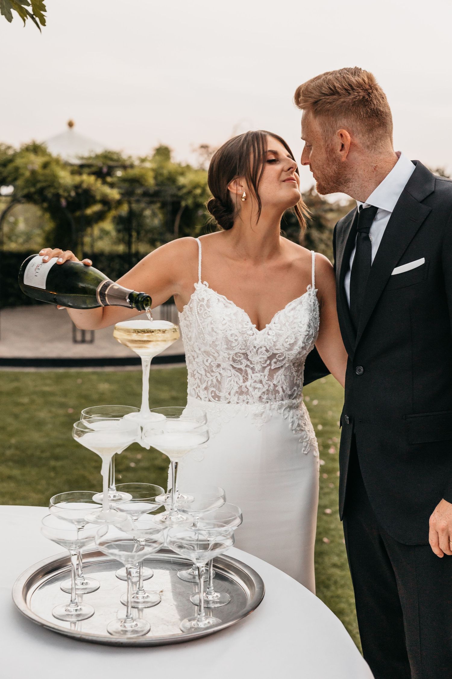 Bride in lace gown pouring champagne tower while groom in black suit watches.