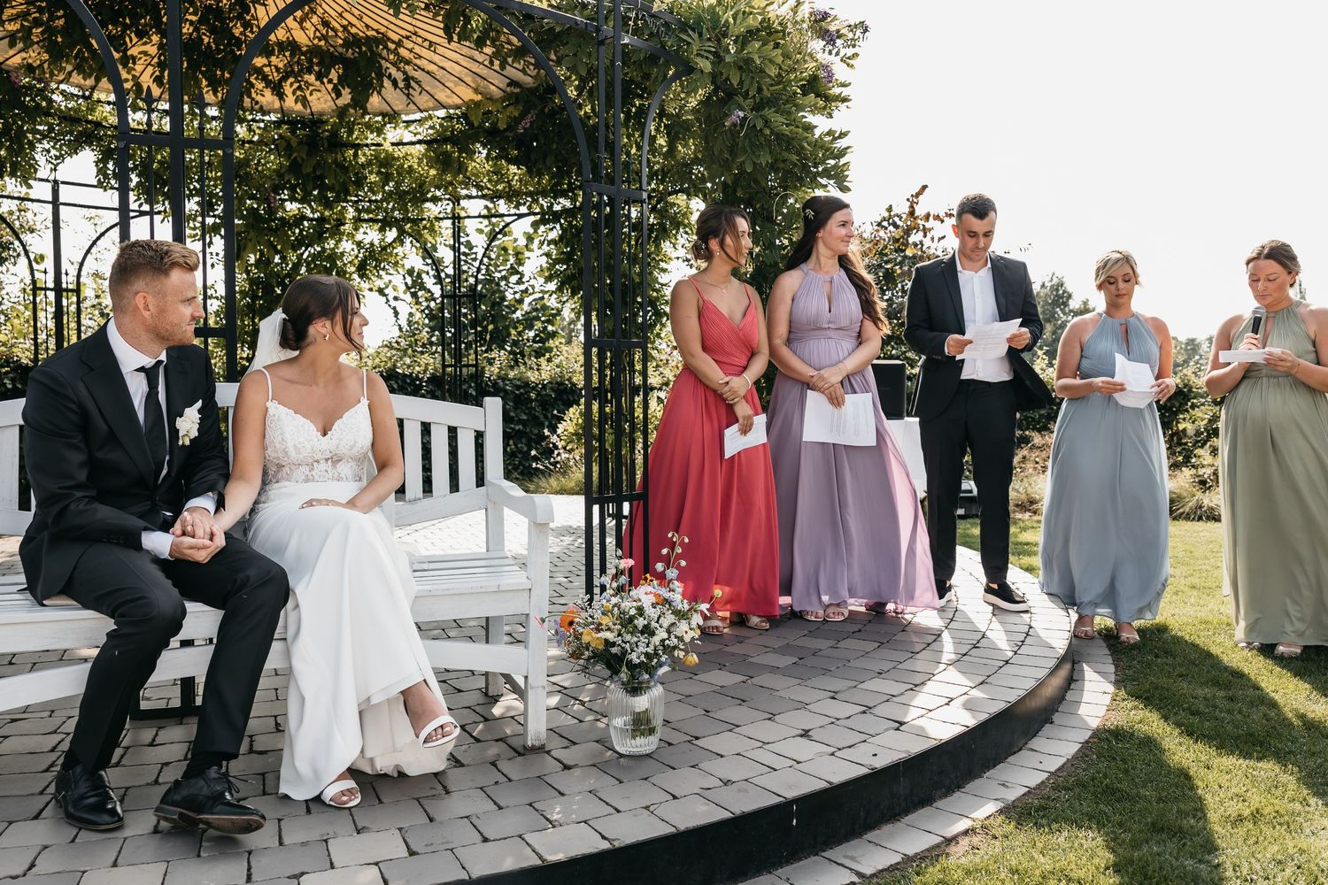 Bride and groom on white bench listening to wedding party readings during outdoor garden ceremony.