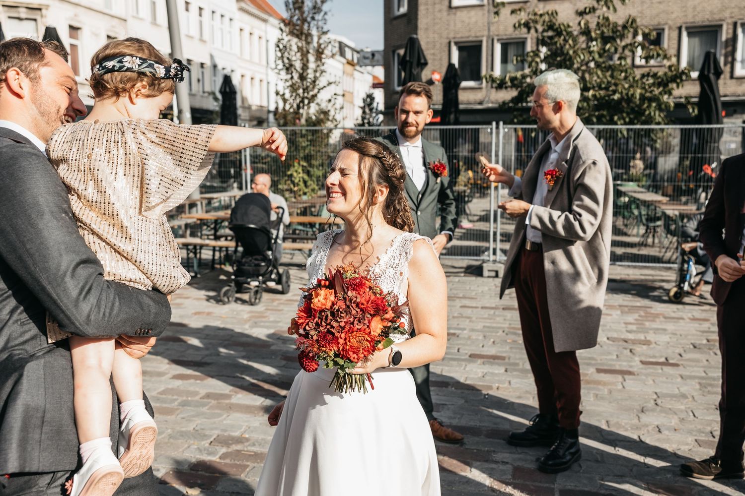 Smiling bride holding red bouquet outdoors at wedding celebration with guests on cobblestone square.