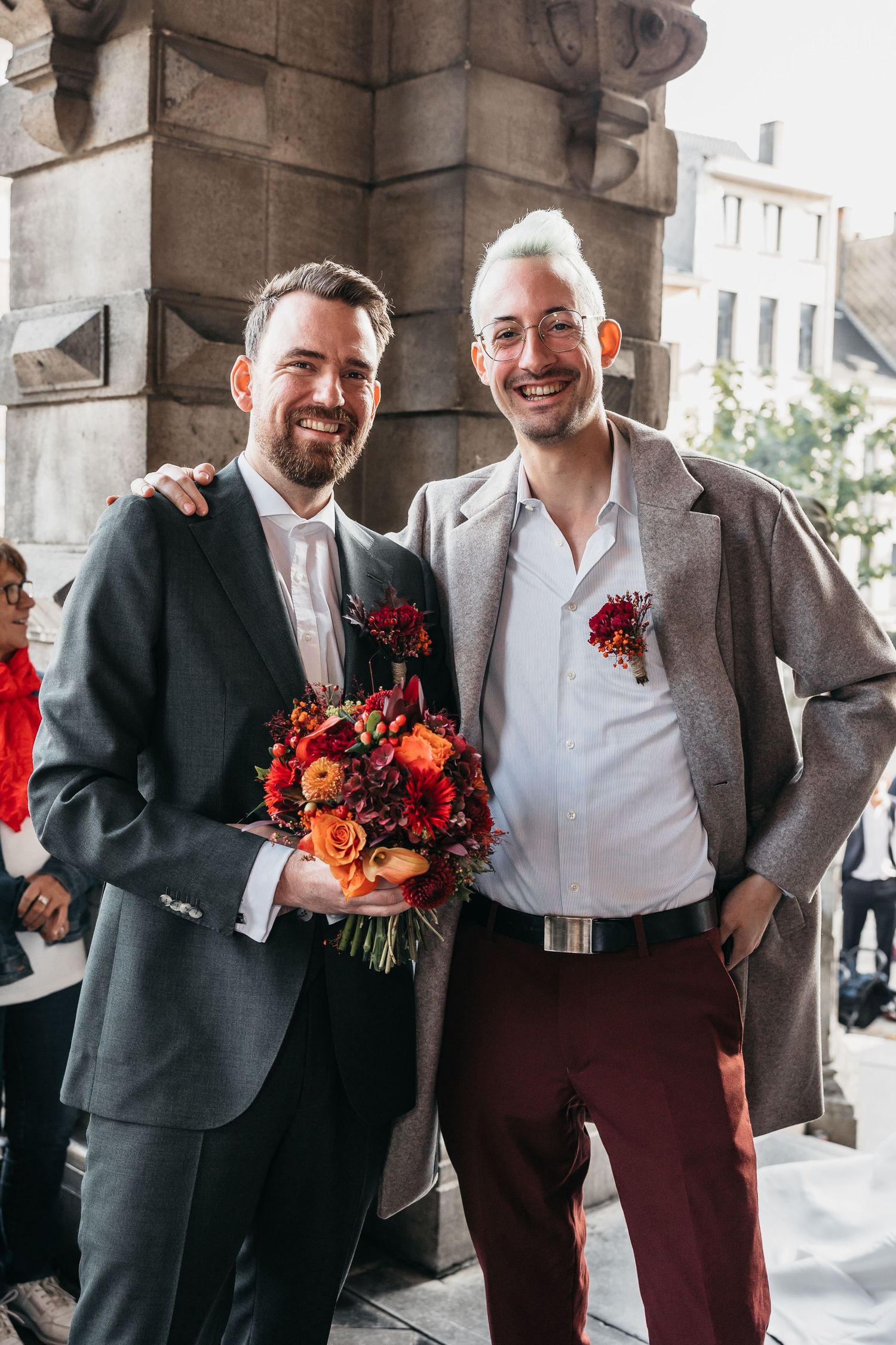 Two smiling men pose together at wedding, one holding colorful red and orange floral bouquet.
