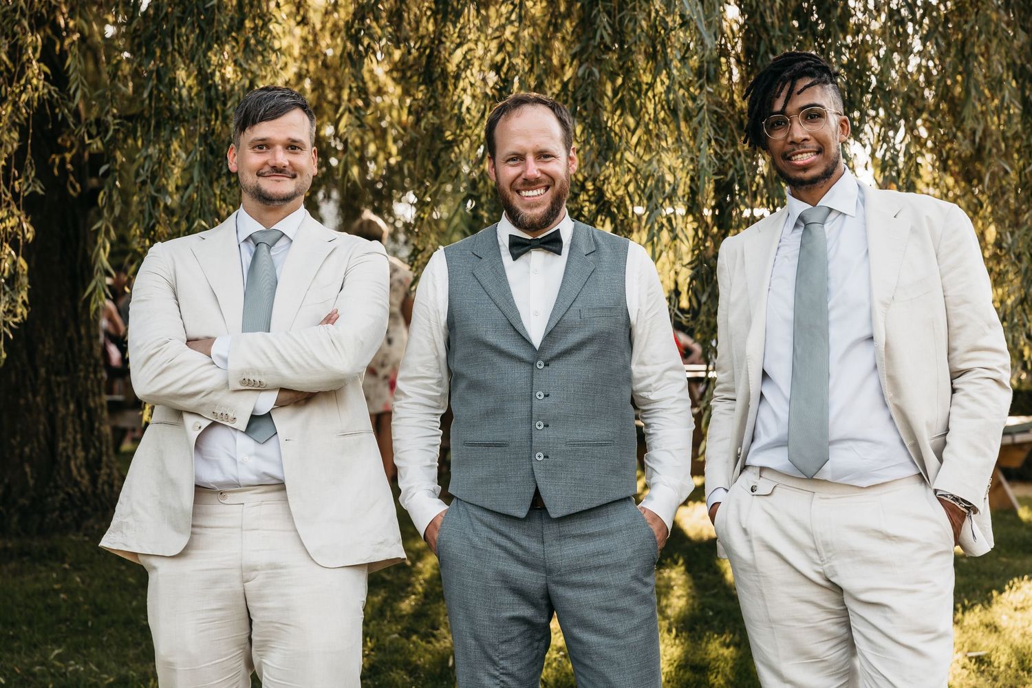 Groom in gray waistcoat and bow tie smiling with two groomsmen in matching cream suits outdoors.