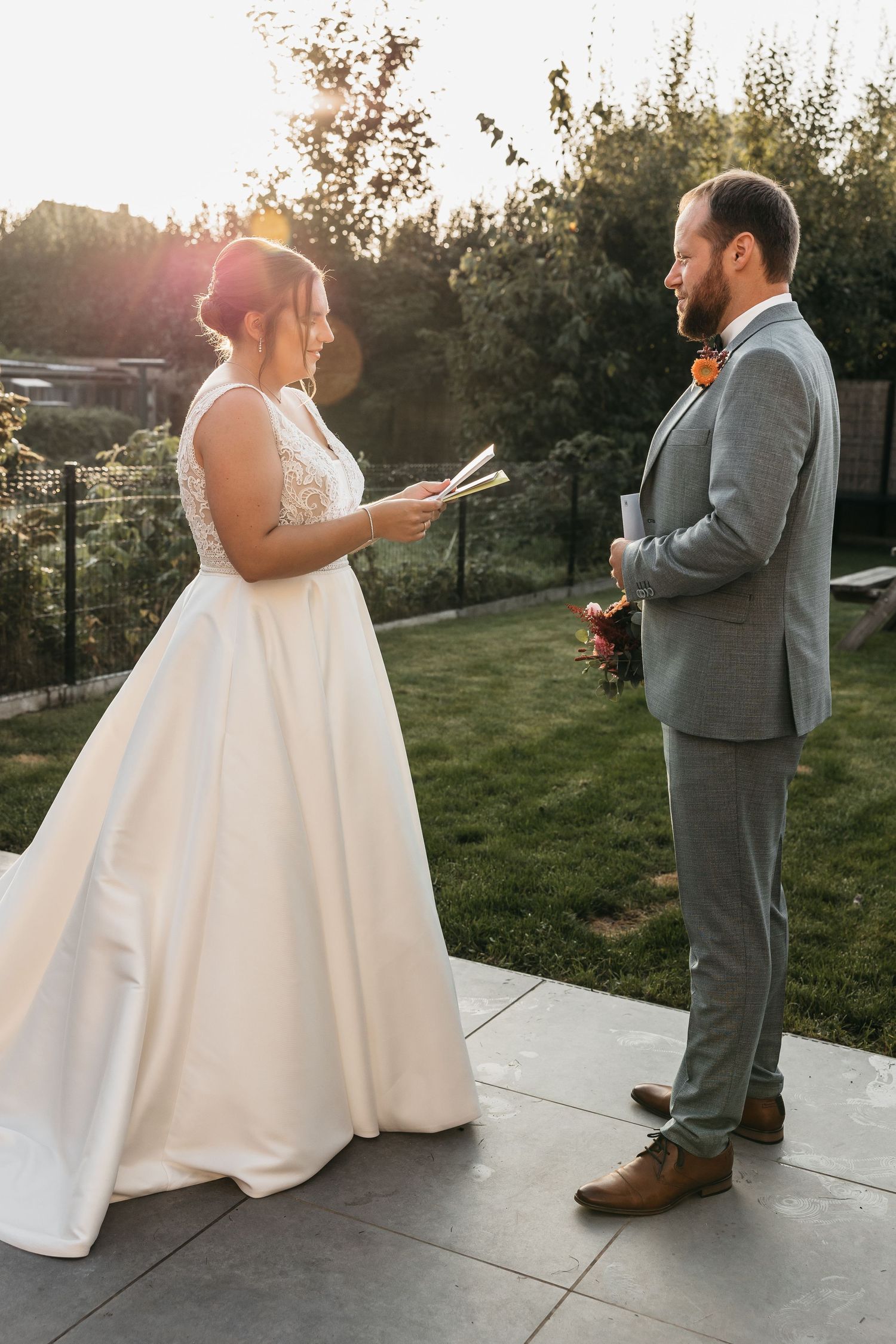 Bride reads private vows from paper to smiling groom in gray suit during outdoor sunset vow exchange.
