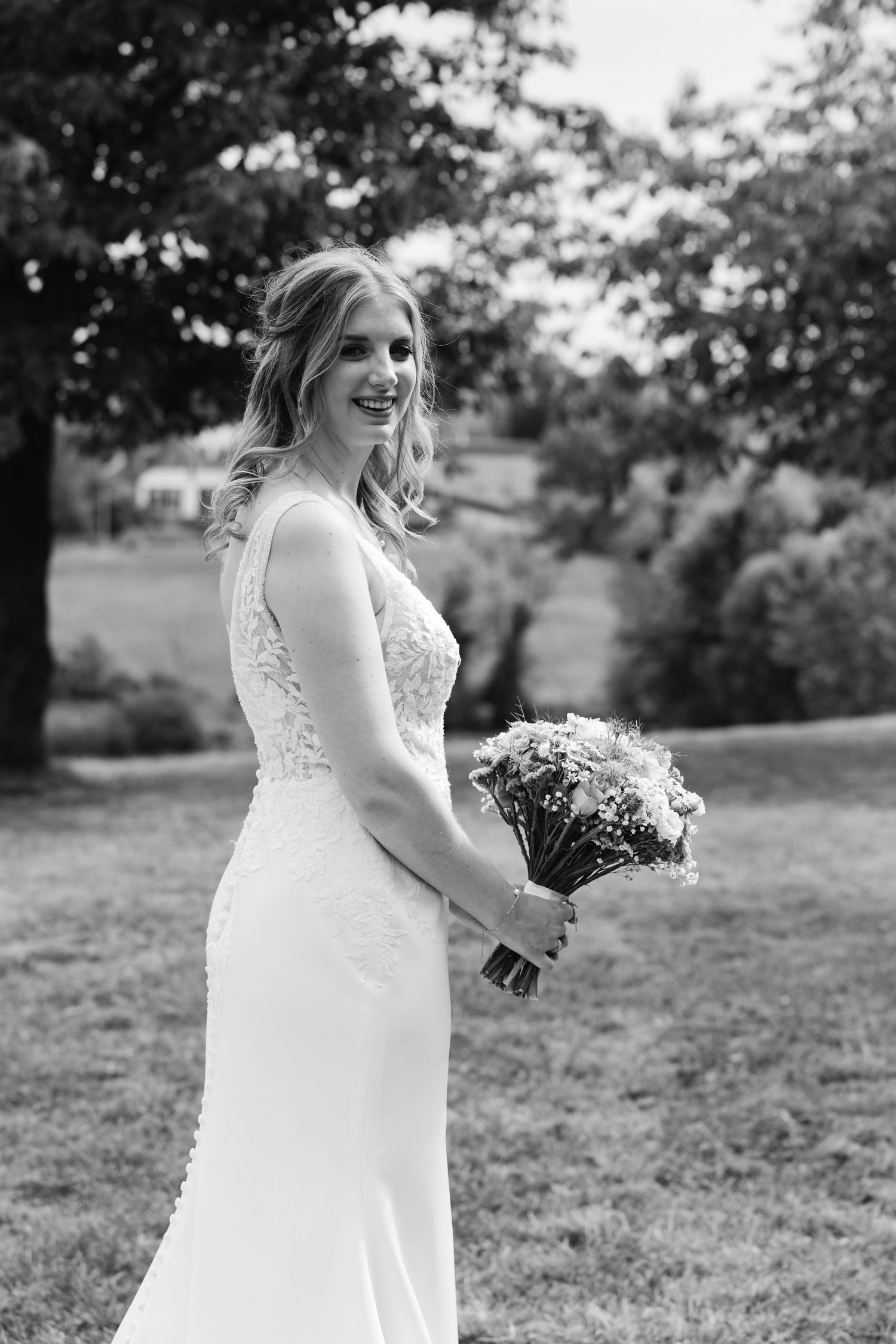 Black and white portrait of a smiling bride in lace gown holding a bouquet in a garden setting.