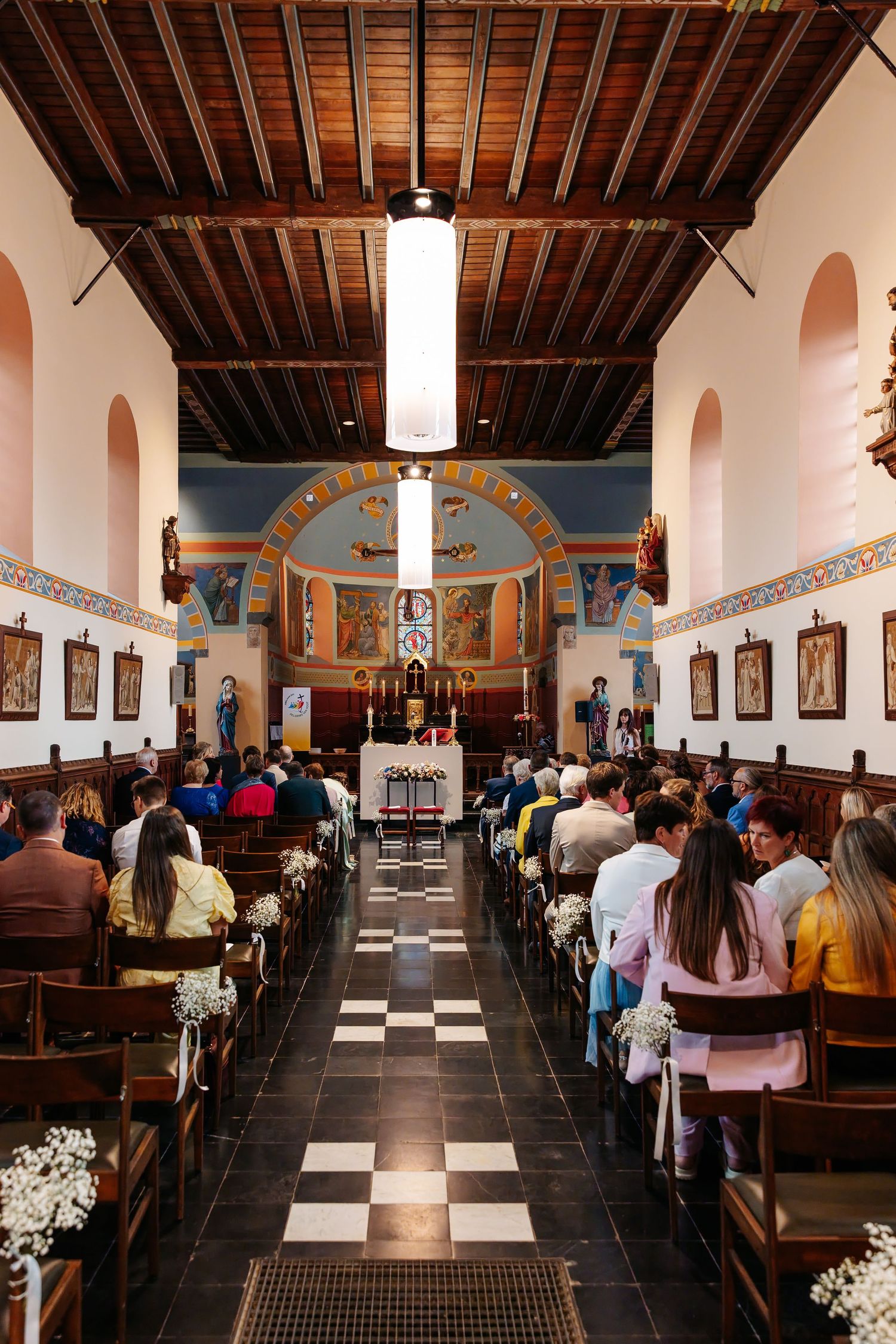 Wedding guests seated in ornate church with colorful murals and checkered floor.