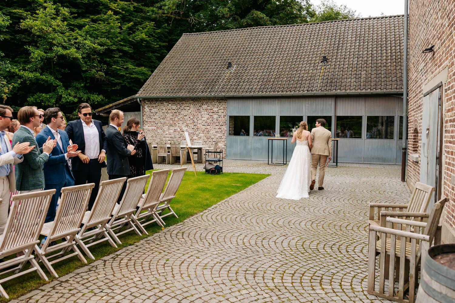 Wedding guests applaud as bride and groom embrace on cobblestone courtyard outside rustic brick venue.