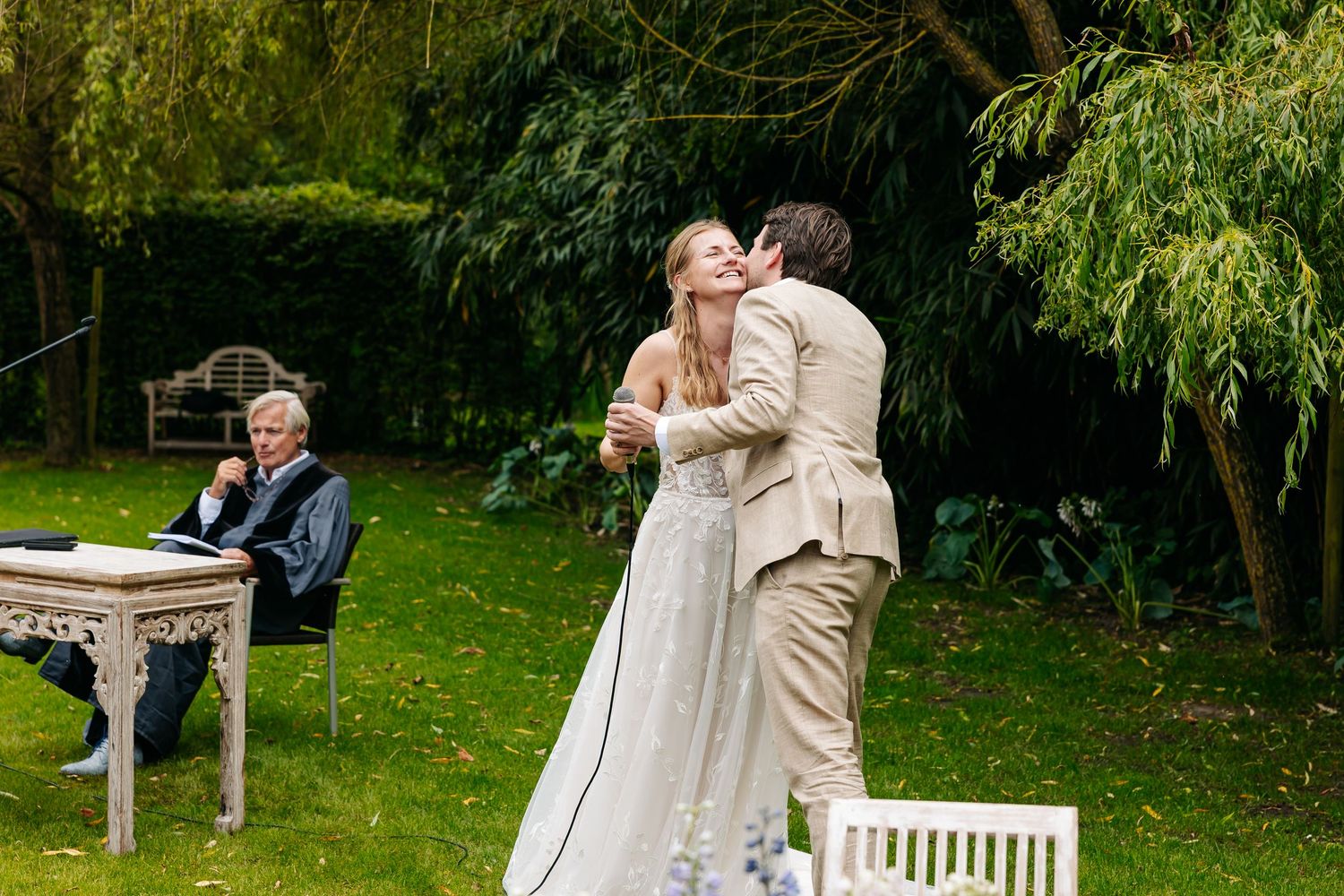 Groom kisses bride during outdoor wedding ceremony in a lush green garden setting.