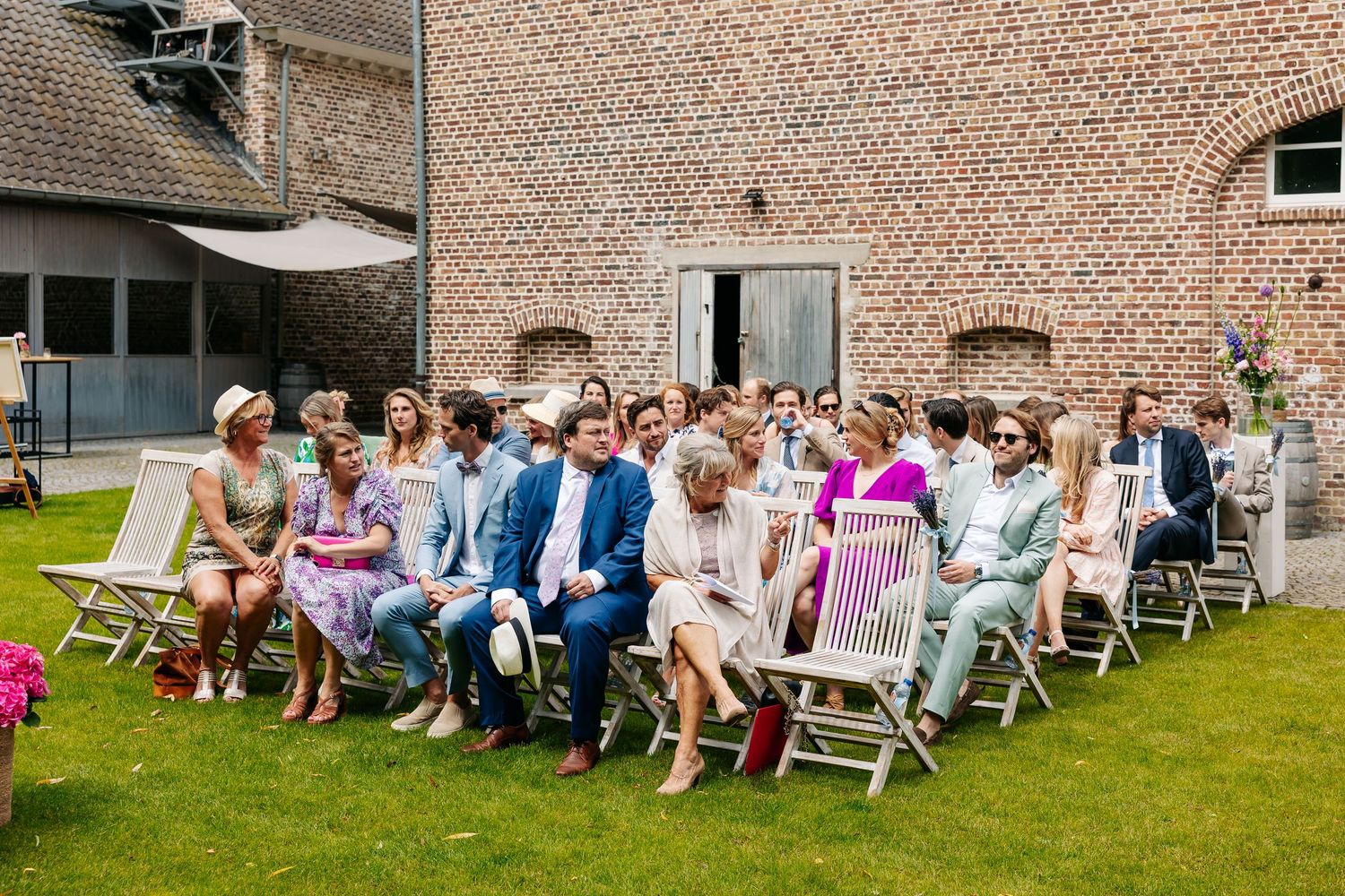 Wedding guests seated on garden chairs at outdoor ceremony beside rustic brick building.