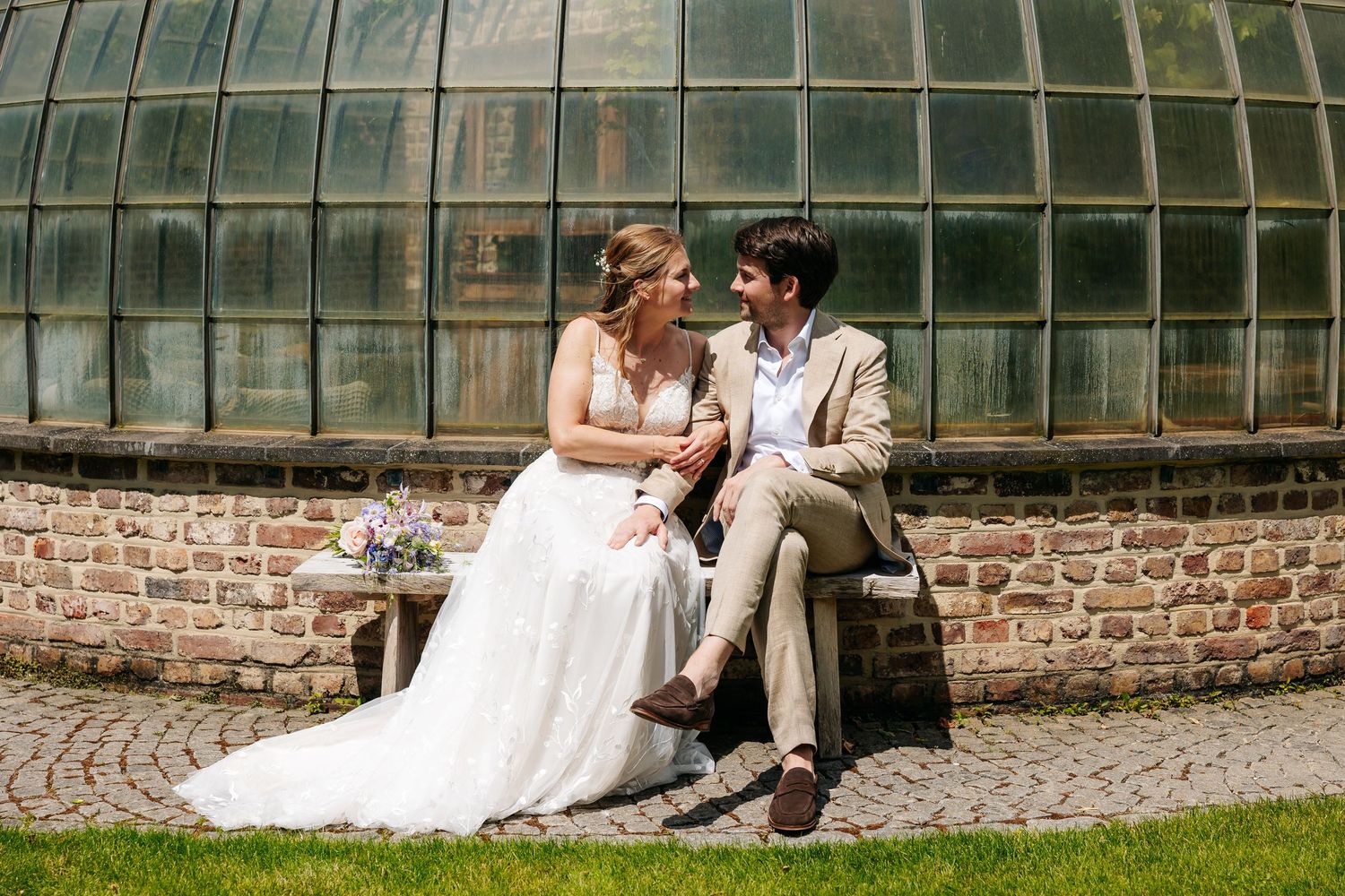 Bride and groom sitting on a stone bench in front of a glass greenhouse, sharing an intimate moment on their wedding day.