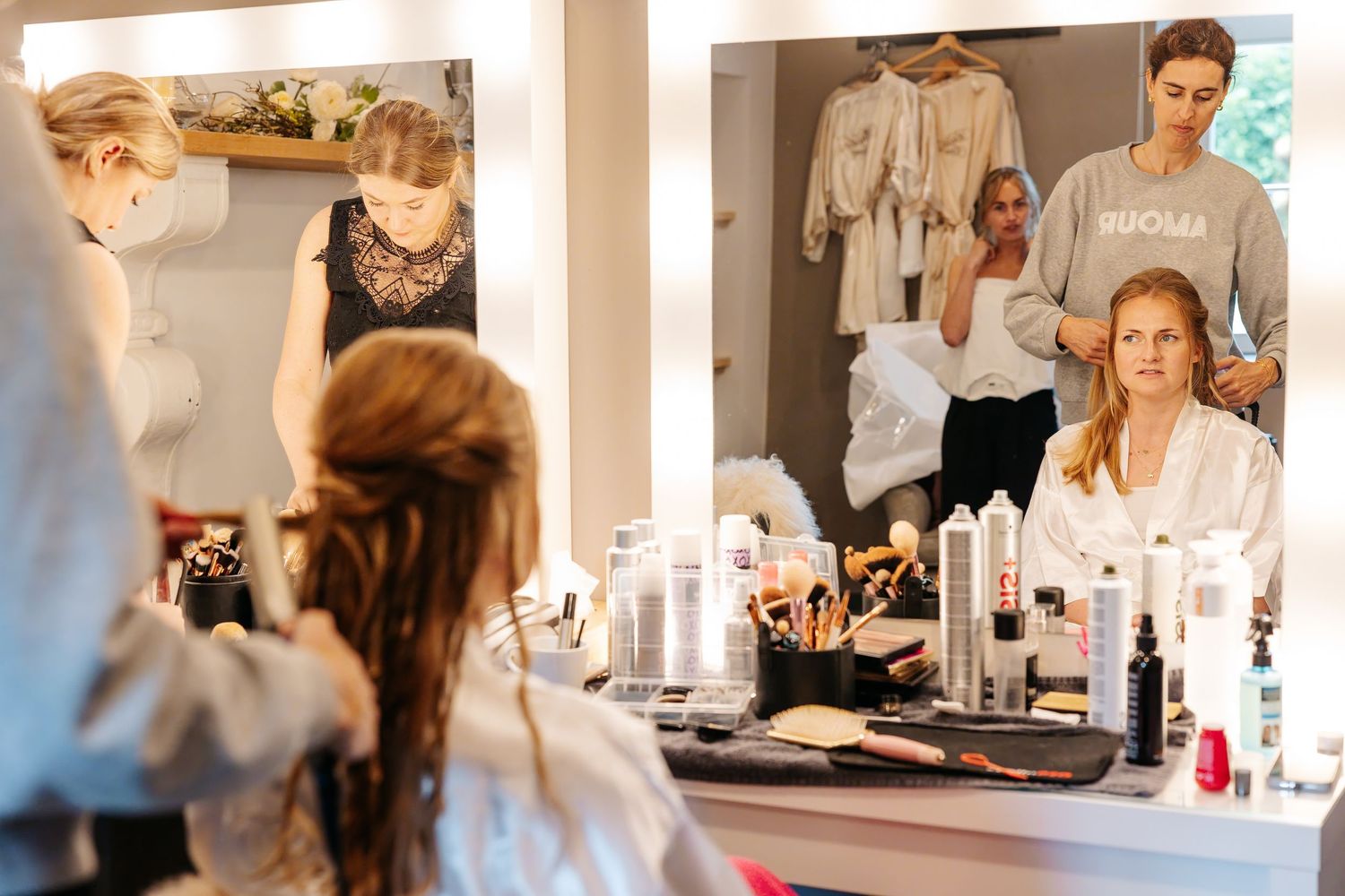Bride and bridesmaids in robes getting ready at a vanity mirror with makeup and hair products.