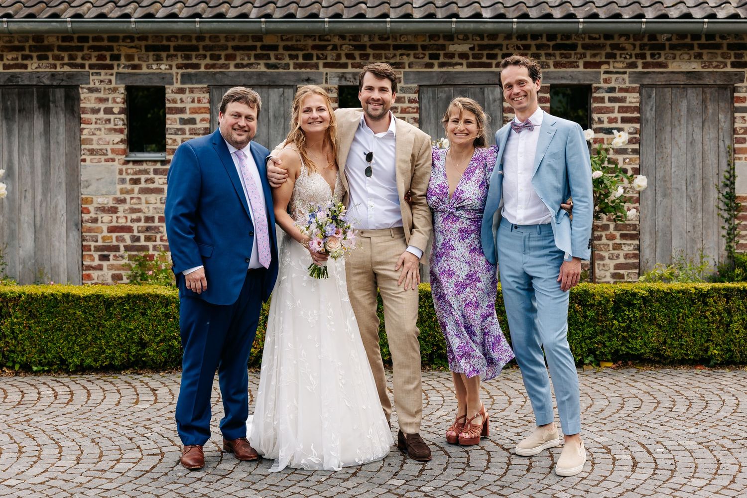 Wedding party of five posing together outdoors at a rustic brick venue, bride in white gown with bouquet.