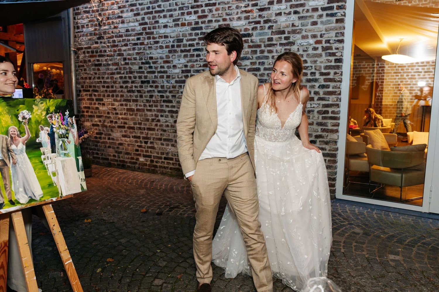 Bride in flowing gown and groom in tan suit smile together outside brick wall at night.