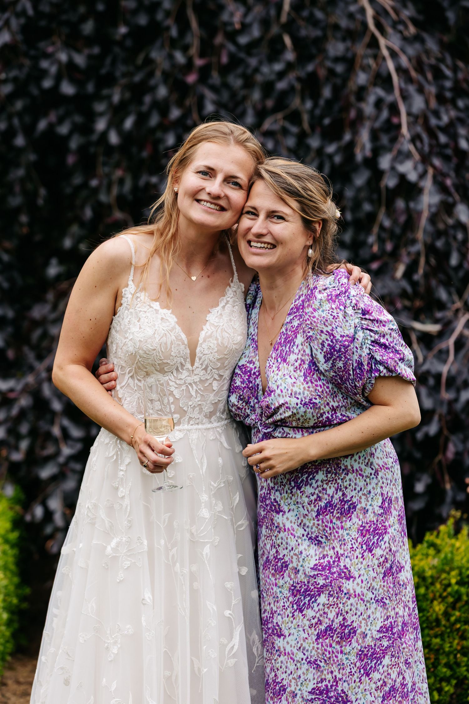 Bride in lace wedding gown smiling with guest in purple floral dress outdoors.