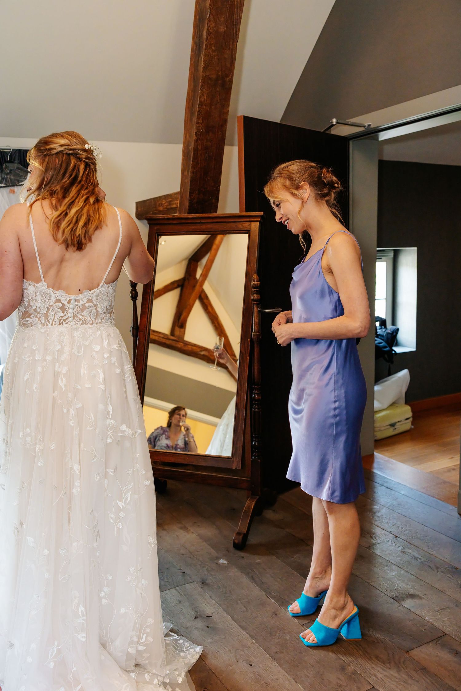 Bridesmaid in blue satin dress helps bride in lace wedding gown prepare in front of a standing mirror.