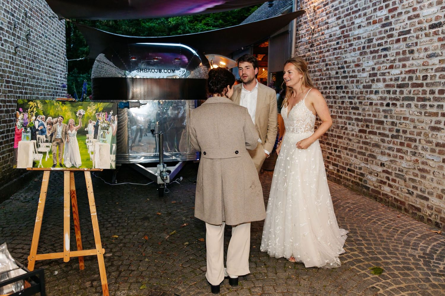 Bride and groom viewing a live wedding painting on an easel outside a rustic brick venue at night.