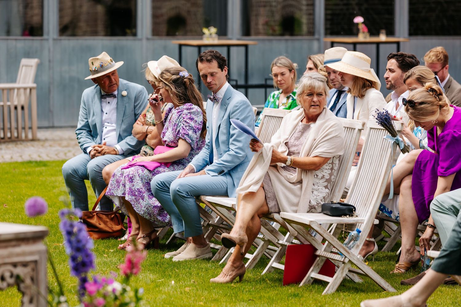 Wedding guests seated on white chairs outdoors during sunny garden ceremony with purple flowers.