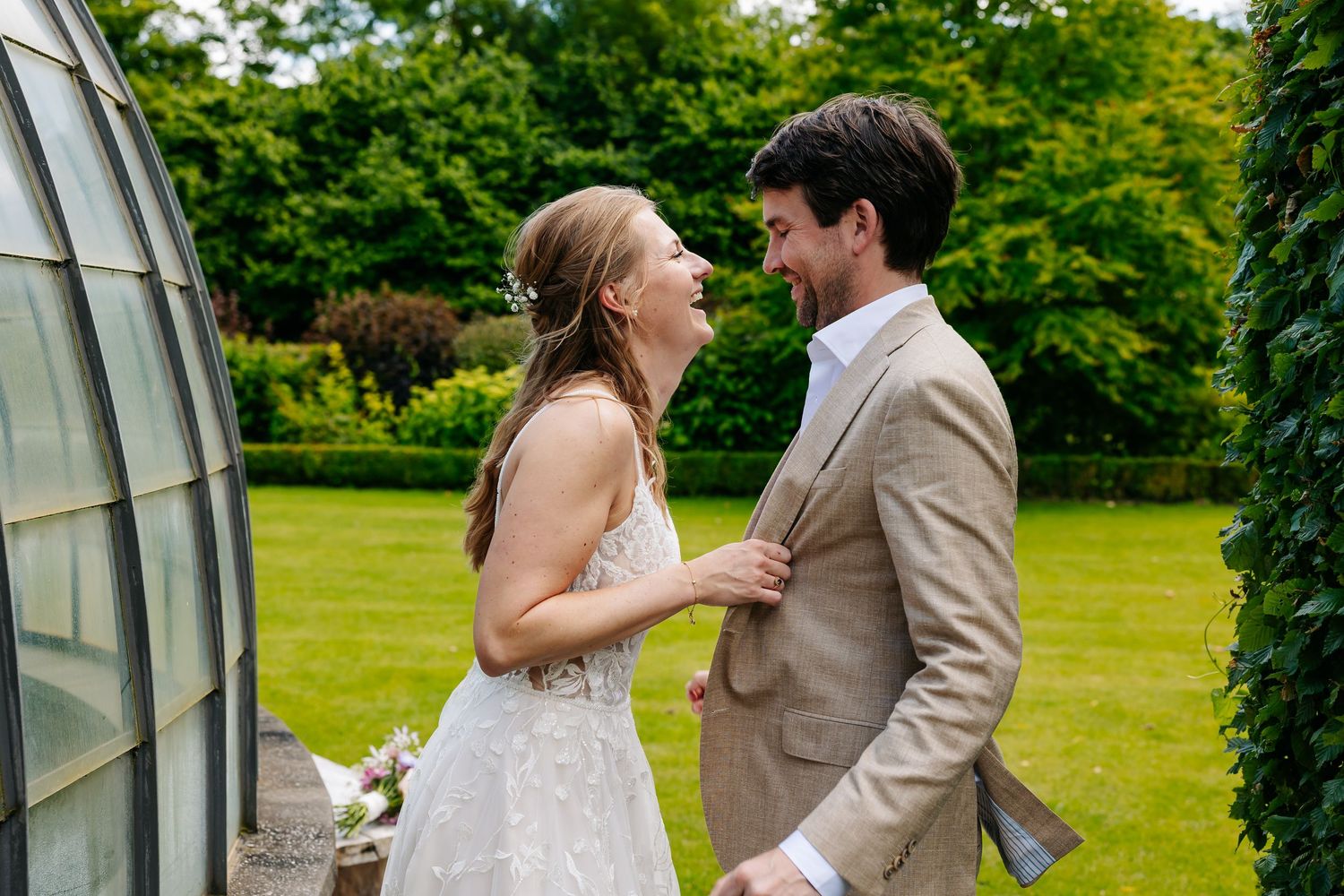 Laughing bride and groom share a joyful moment outdoors near a greenhouse on their wedding day.