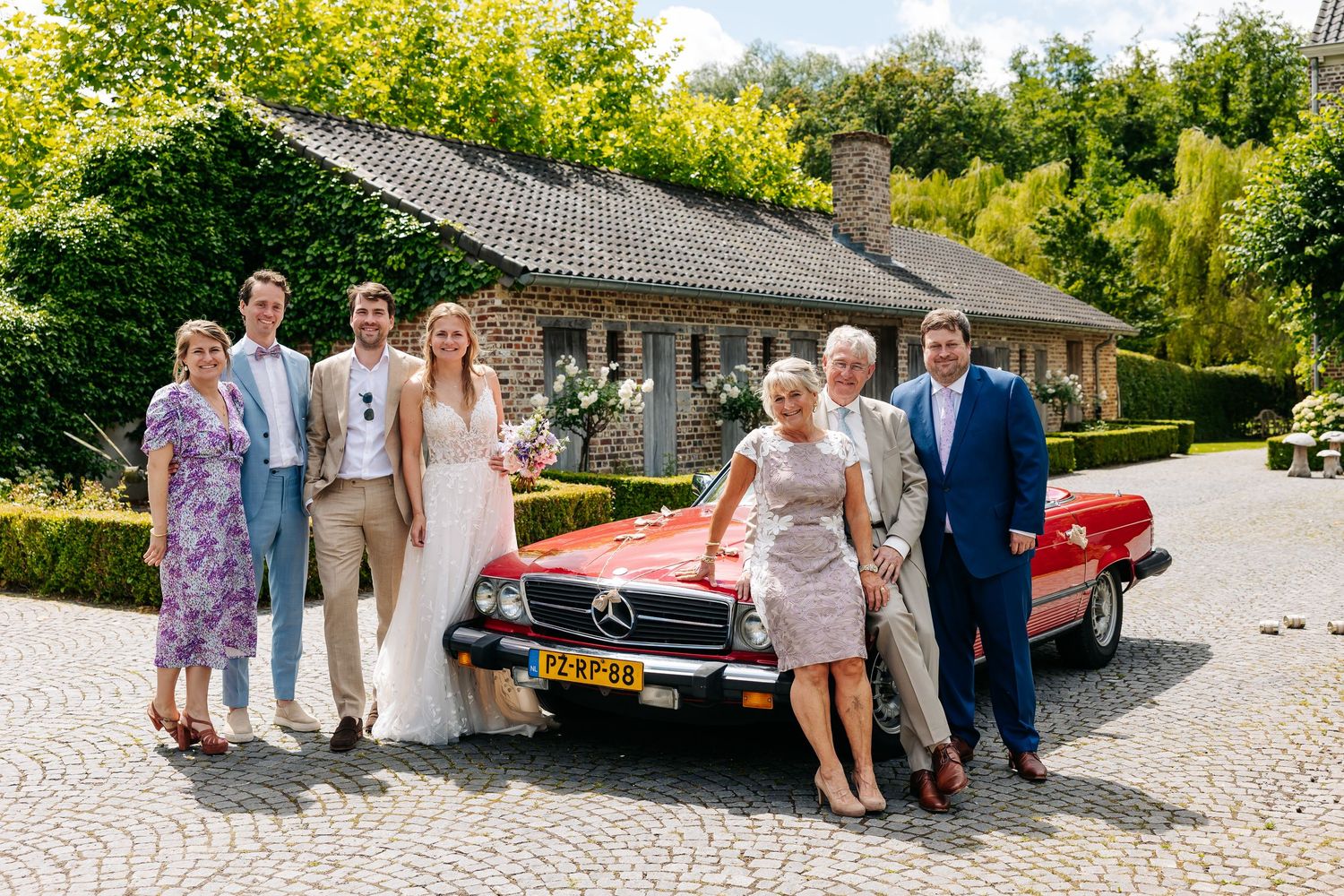 Wedding party posing beside a classic red Mercedes convertible outside a rustic brick farmhouse.