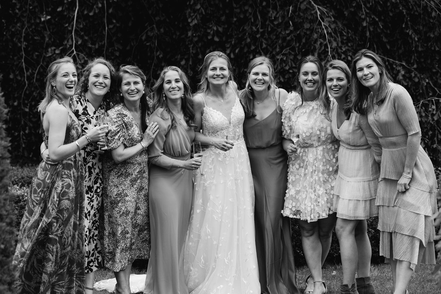Black and white group photo of smiling bride in lace gown with eight bridesmaids outdoors.