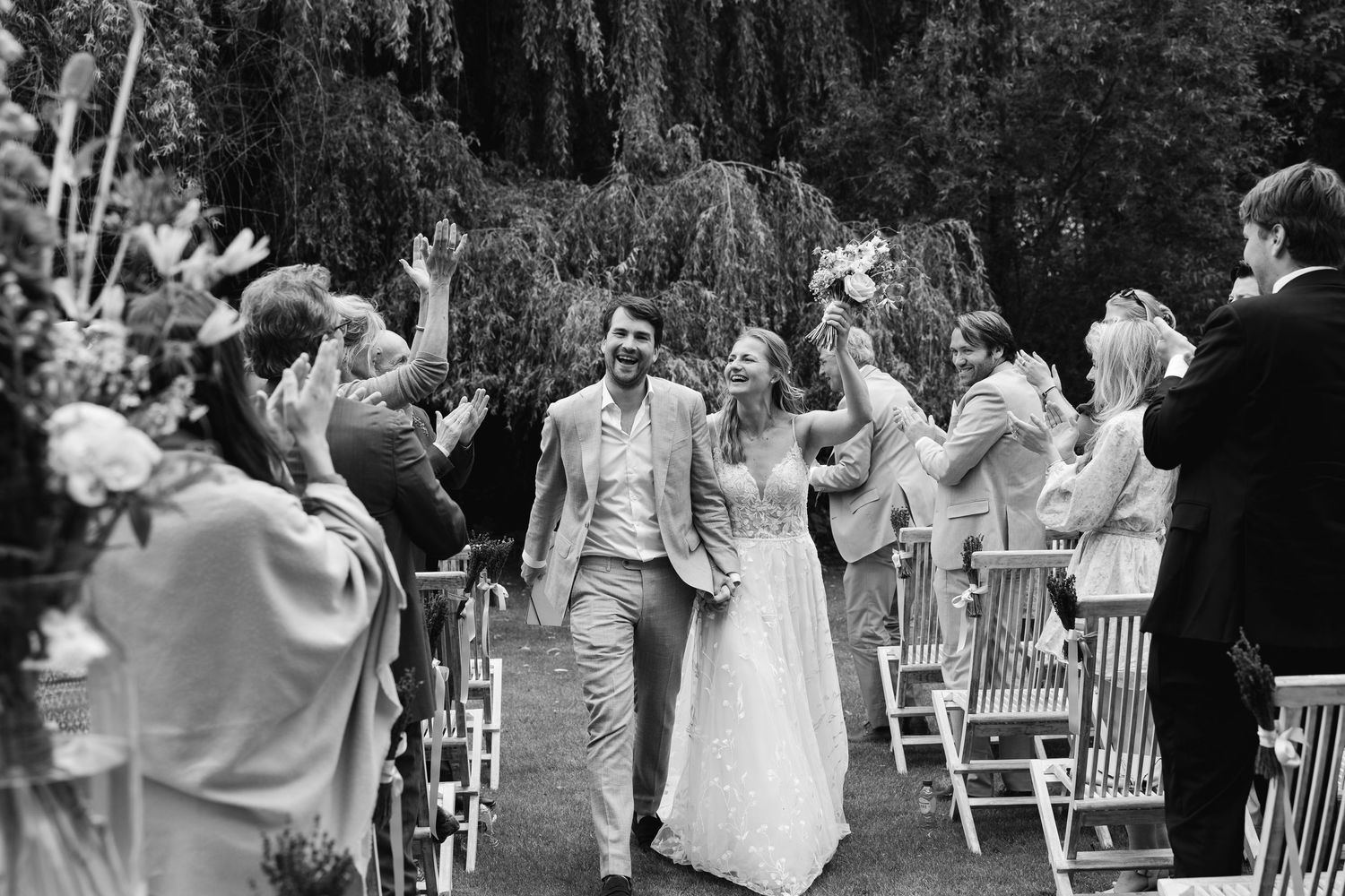 Black and white photo of newlyweds walking down aisle as guests cheer and wave at garden wedding.