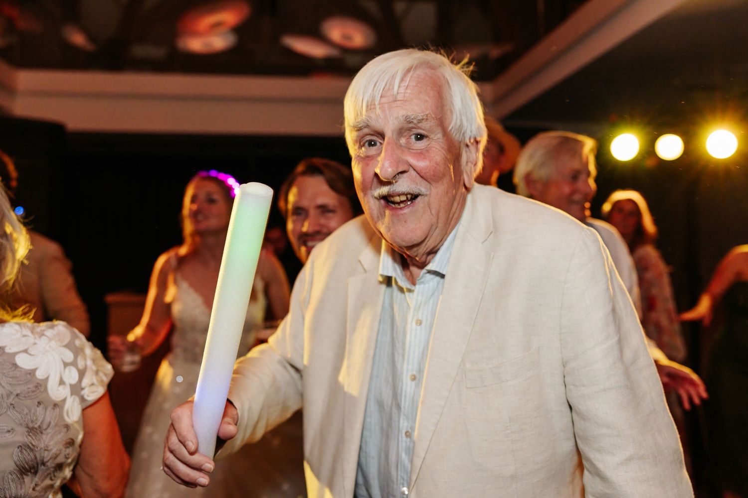 Elderly man smiling and holding a glowing foam stick at a lively wedding reception.