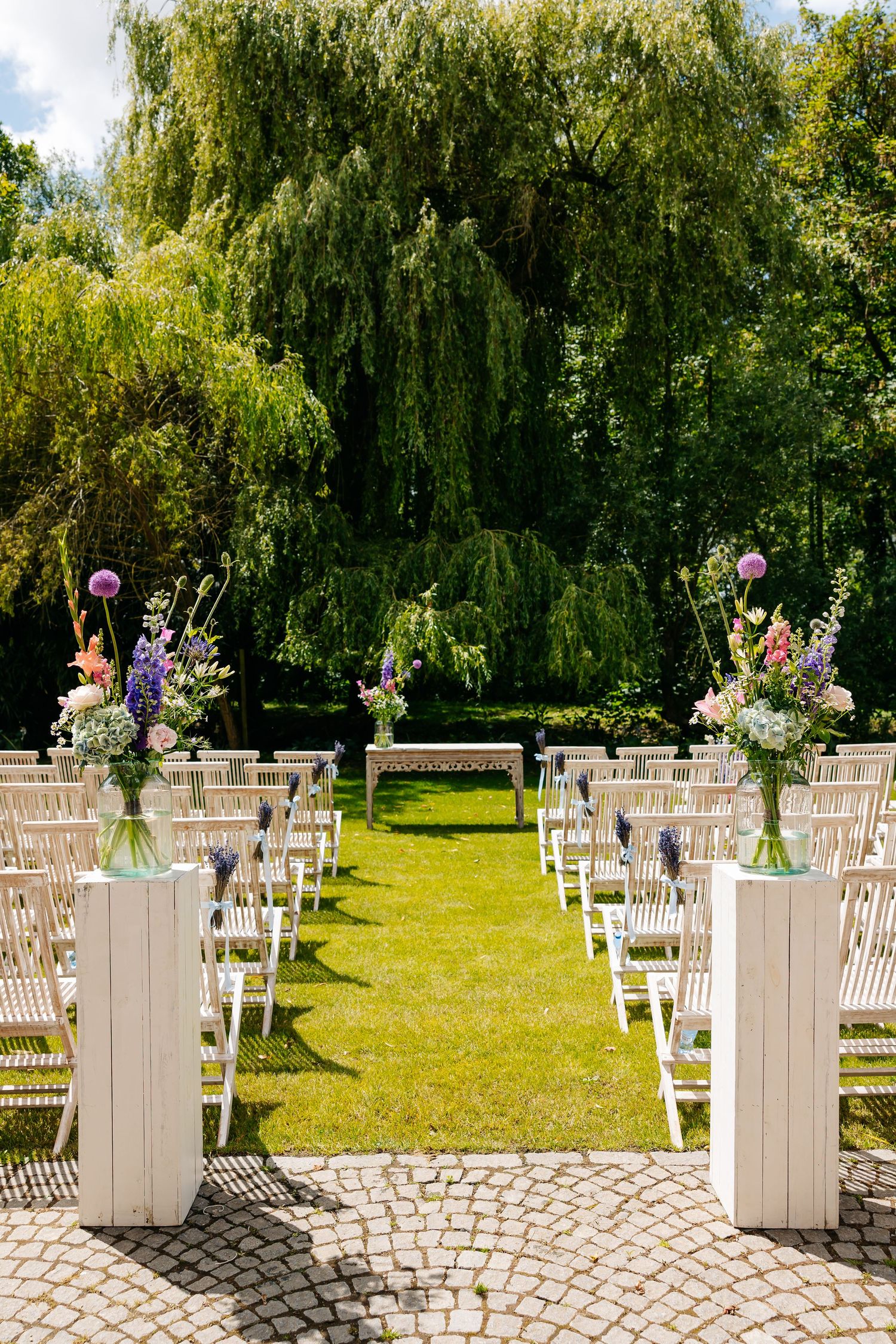 Outdoor wedding ceremony aisle with white chairs and colorful floral arrangements under willow tree.