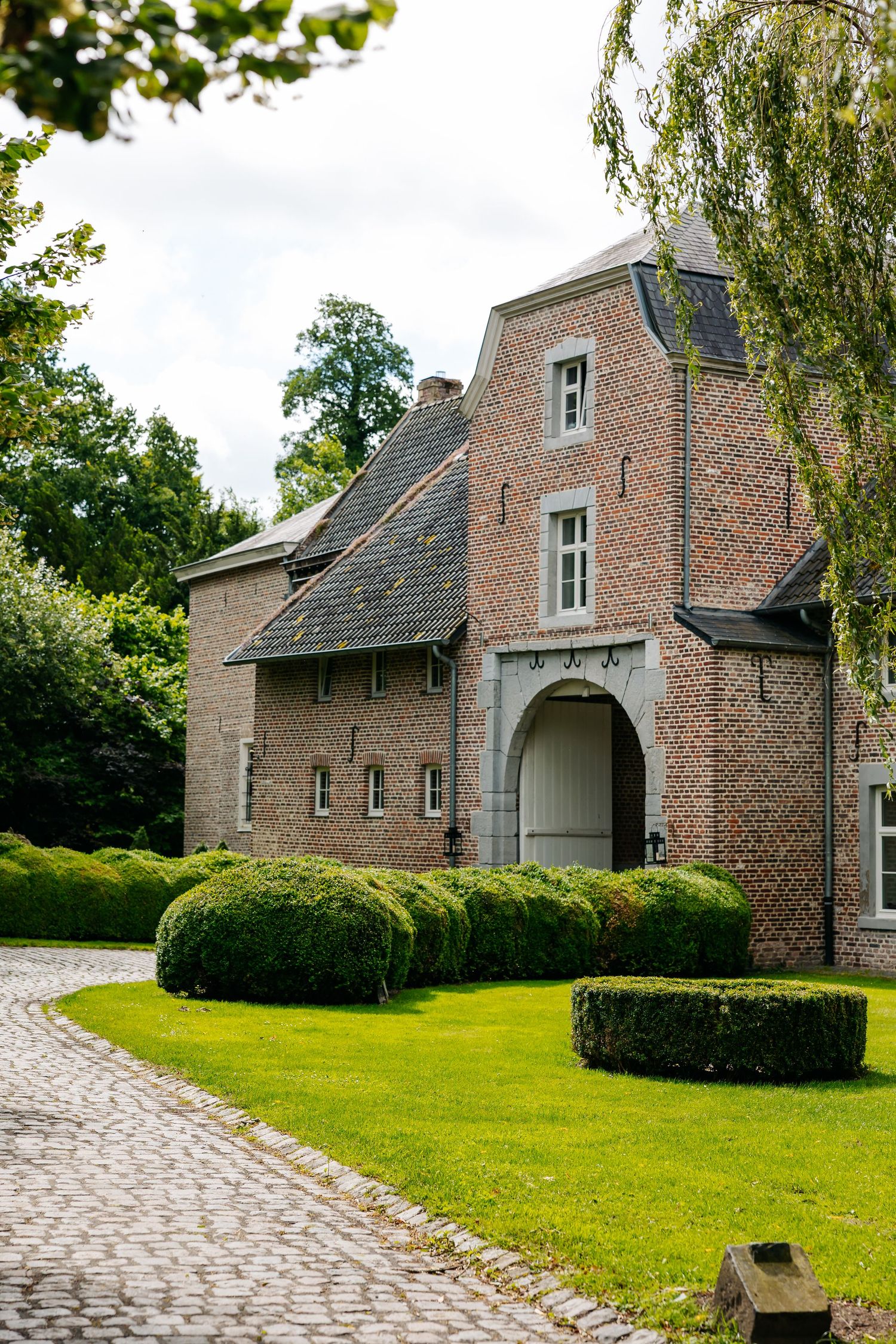 Historic brick manor house with manicured hedges and cobblestone driveway in lush garden.