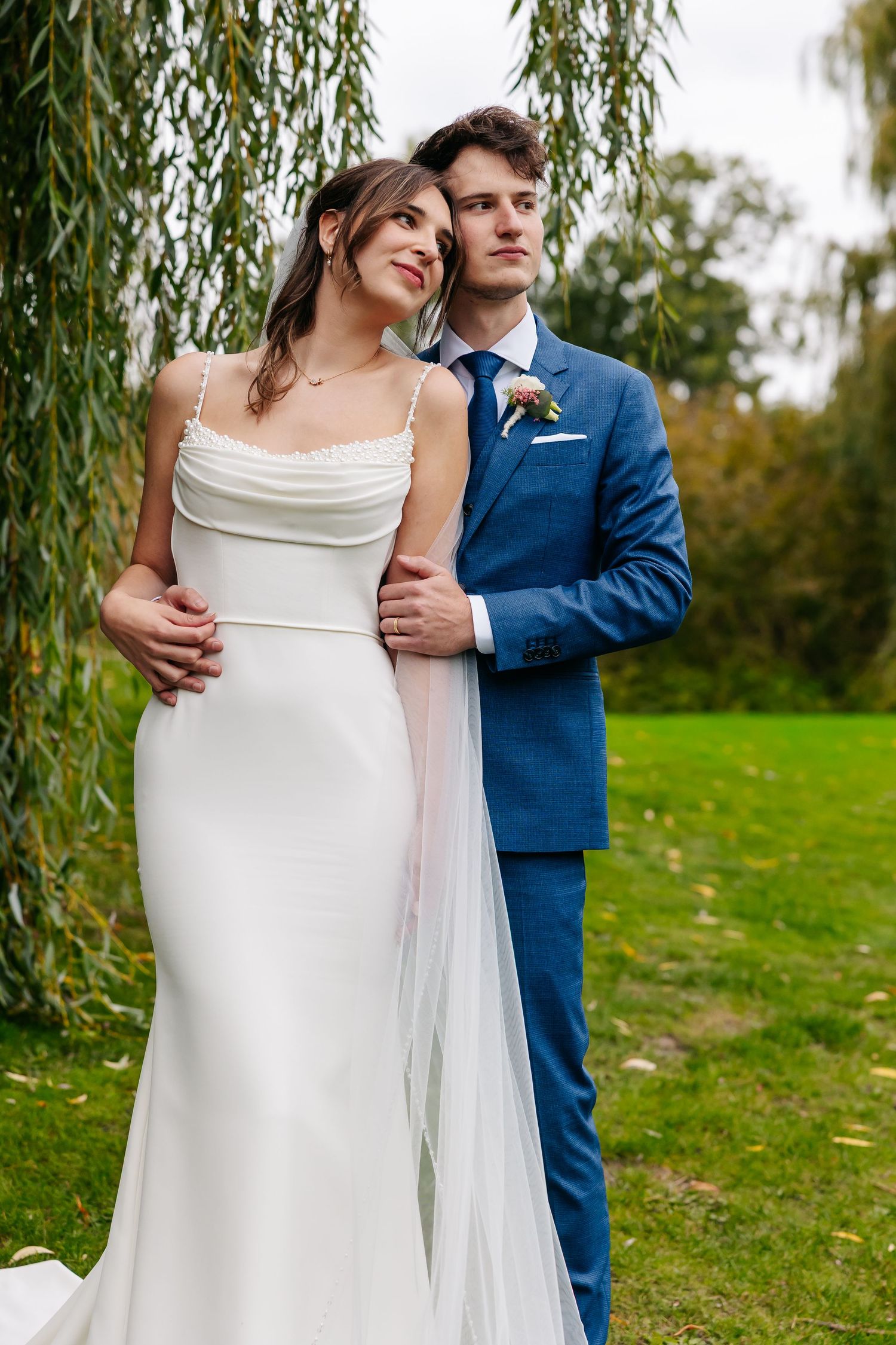 Bride in sleek white dress with veil embraced by groom in blue suit under weeping willow tree.