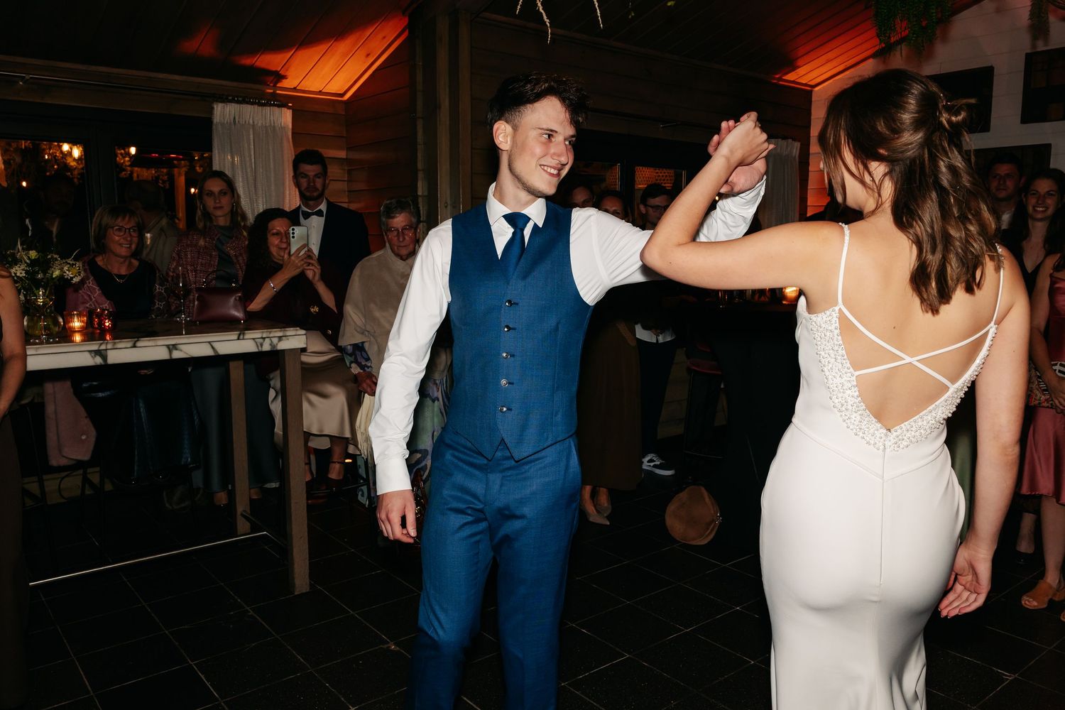 Groom in blue vest dancing with bride in backless white gown at wedding reception.
