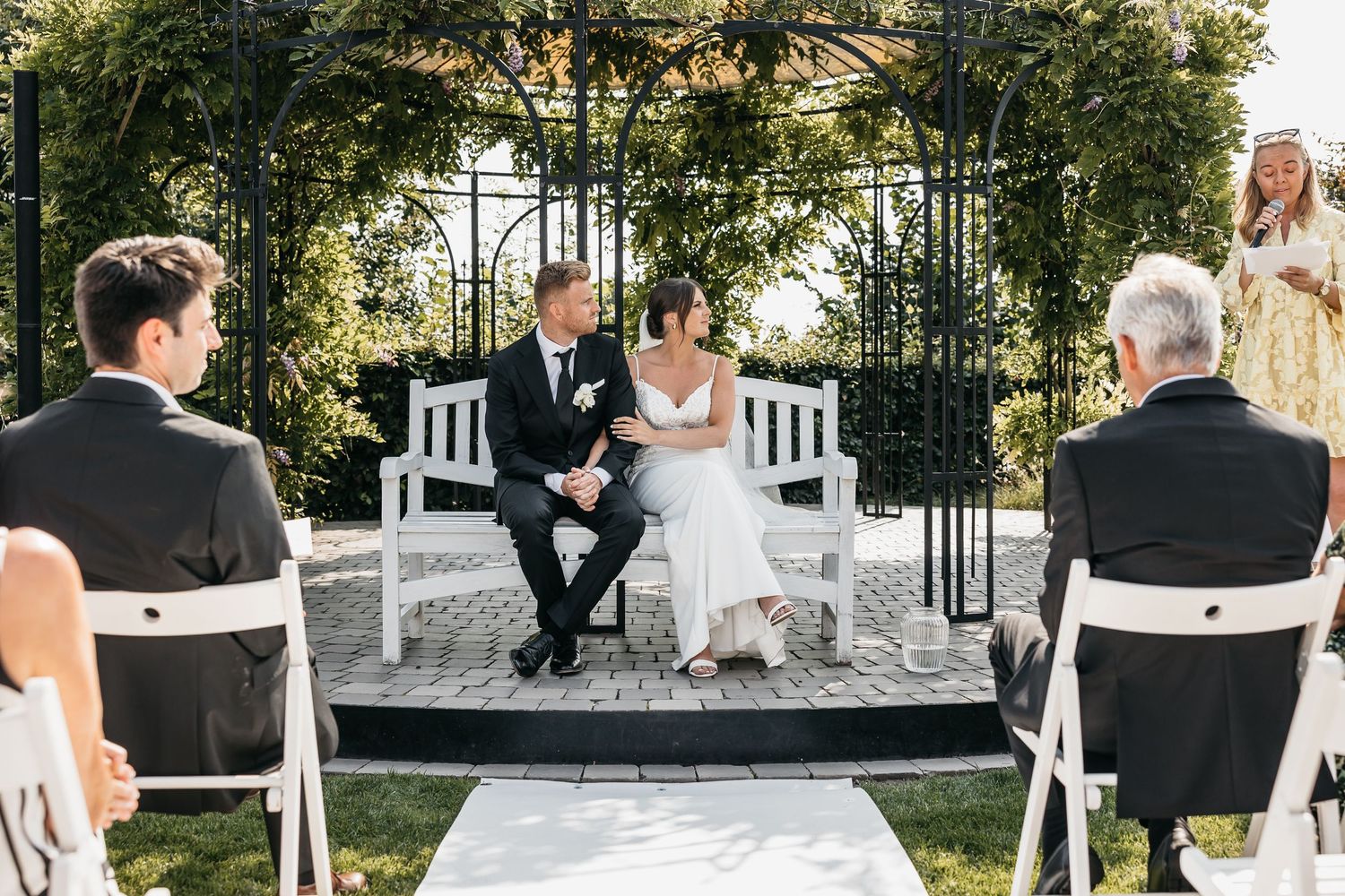 Bride and groom seated on a white bench during outdoor garden wedding ceremony with officiant speaking.
