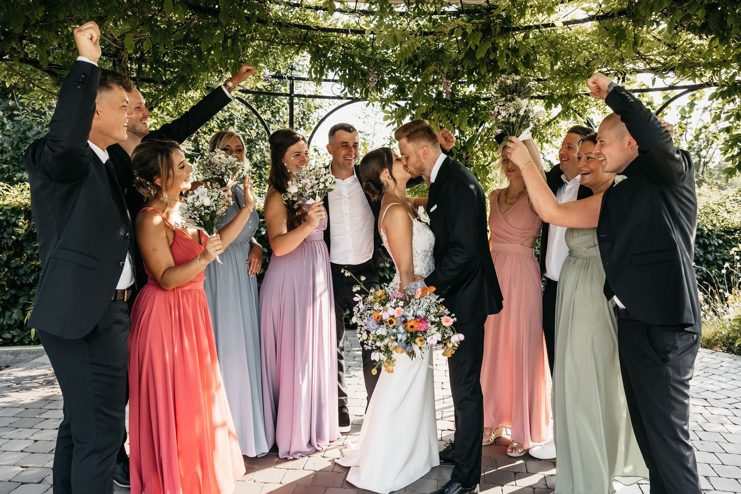 Joyful wedding party cheering as bride and groom kiss beneath a decorative floral arch outdoors.