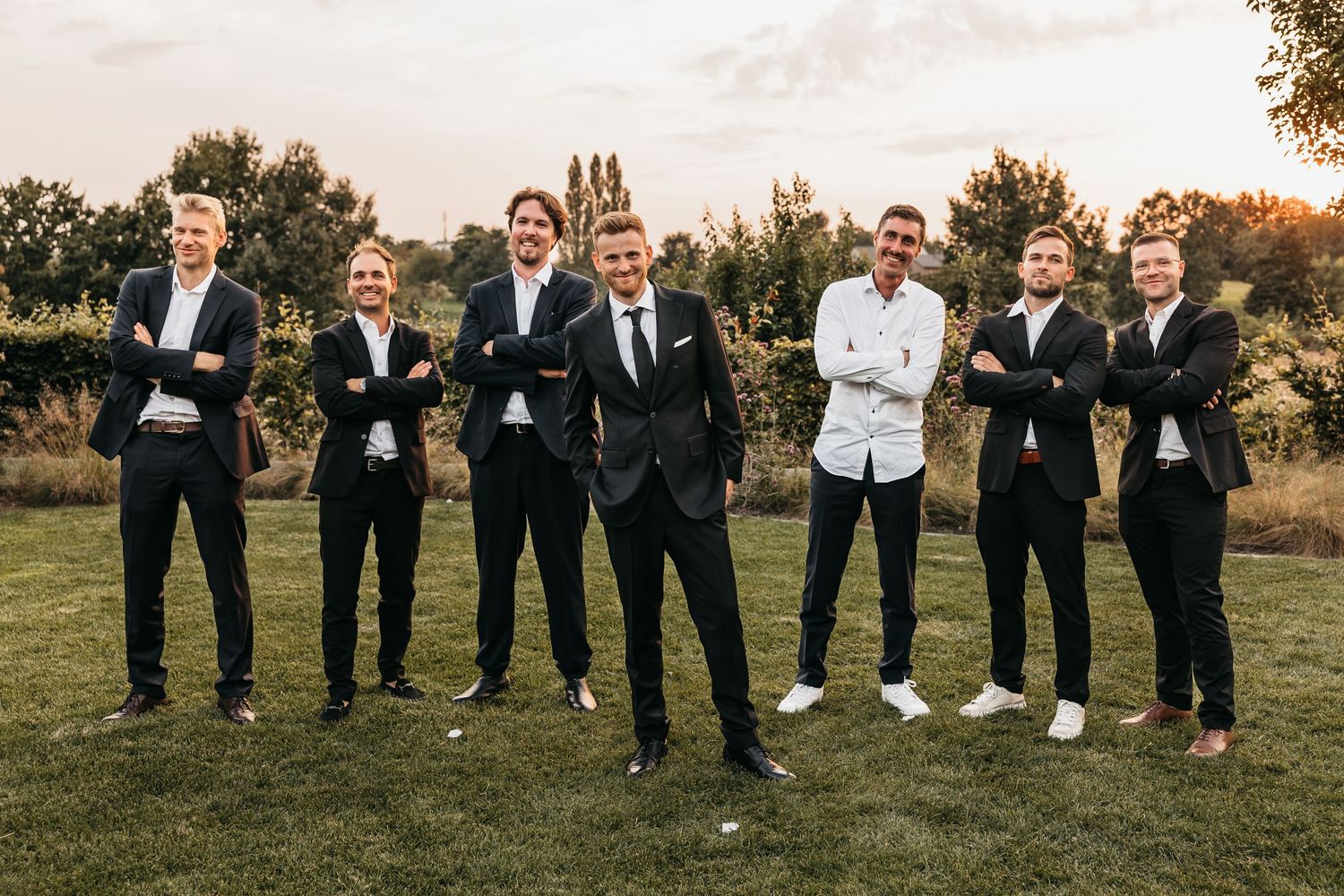 Seven groomsmen in dark suits posing confidently with arms crossed on green lawn.