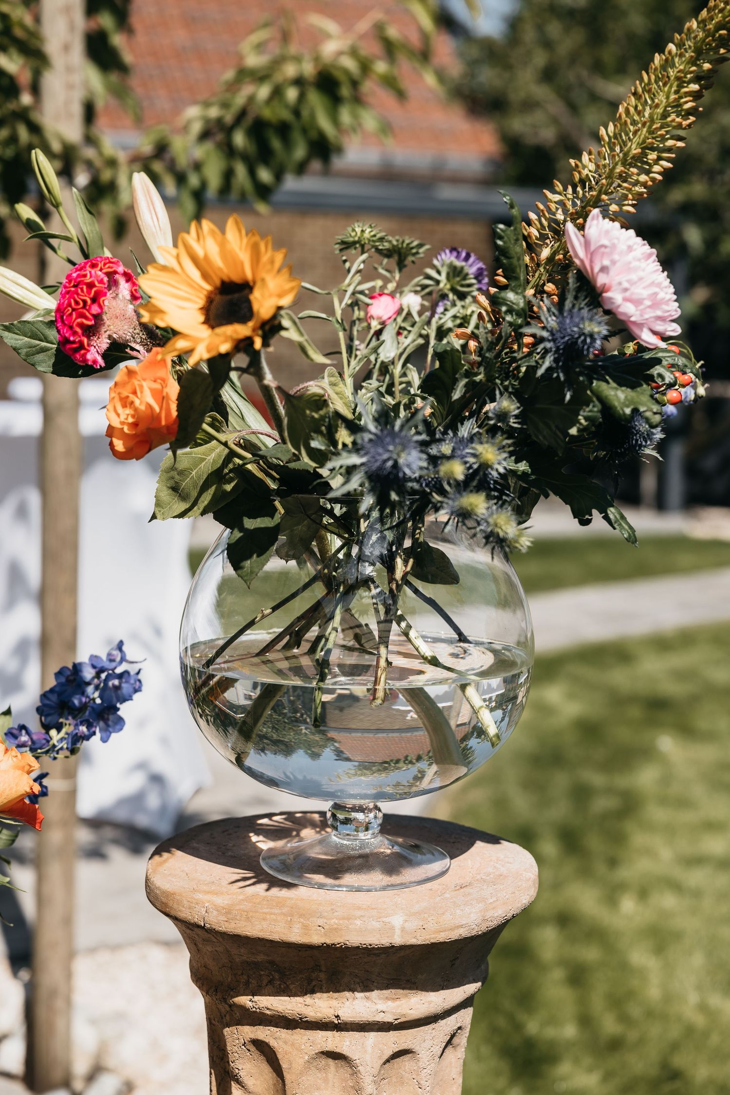Colorful wedding floral arrangement in a round glass vase on a stone pedestal outdoors.