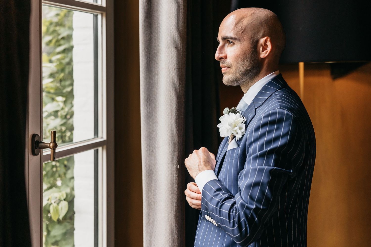 Bald groom in navy pinstripe suit with white boutonniere gazes out window on wedding day.