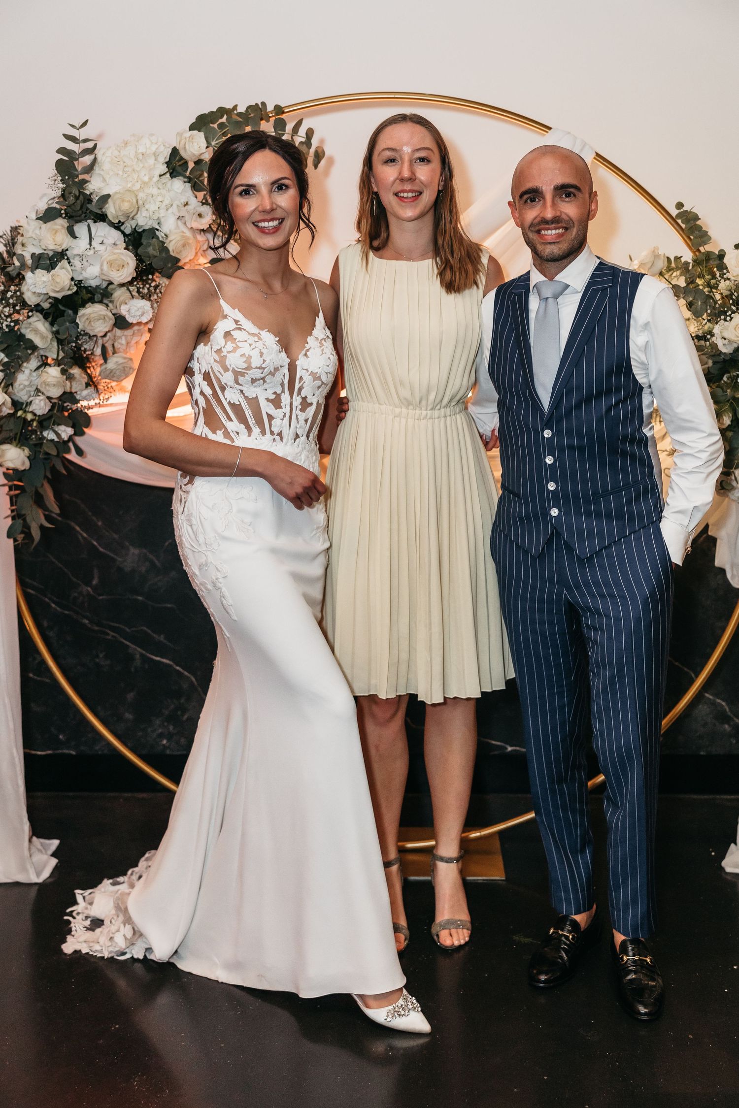 Bride in lace mermaid gown and groom in navy pinstripe suit posing with guest before floral arch backdrop.