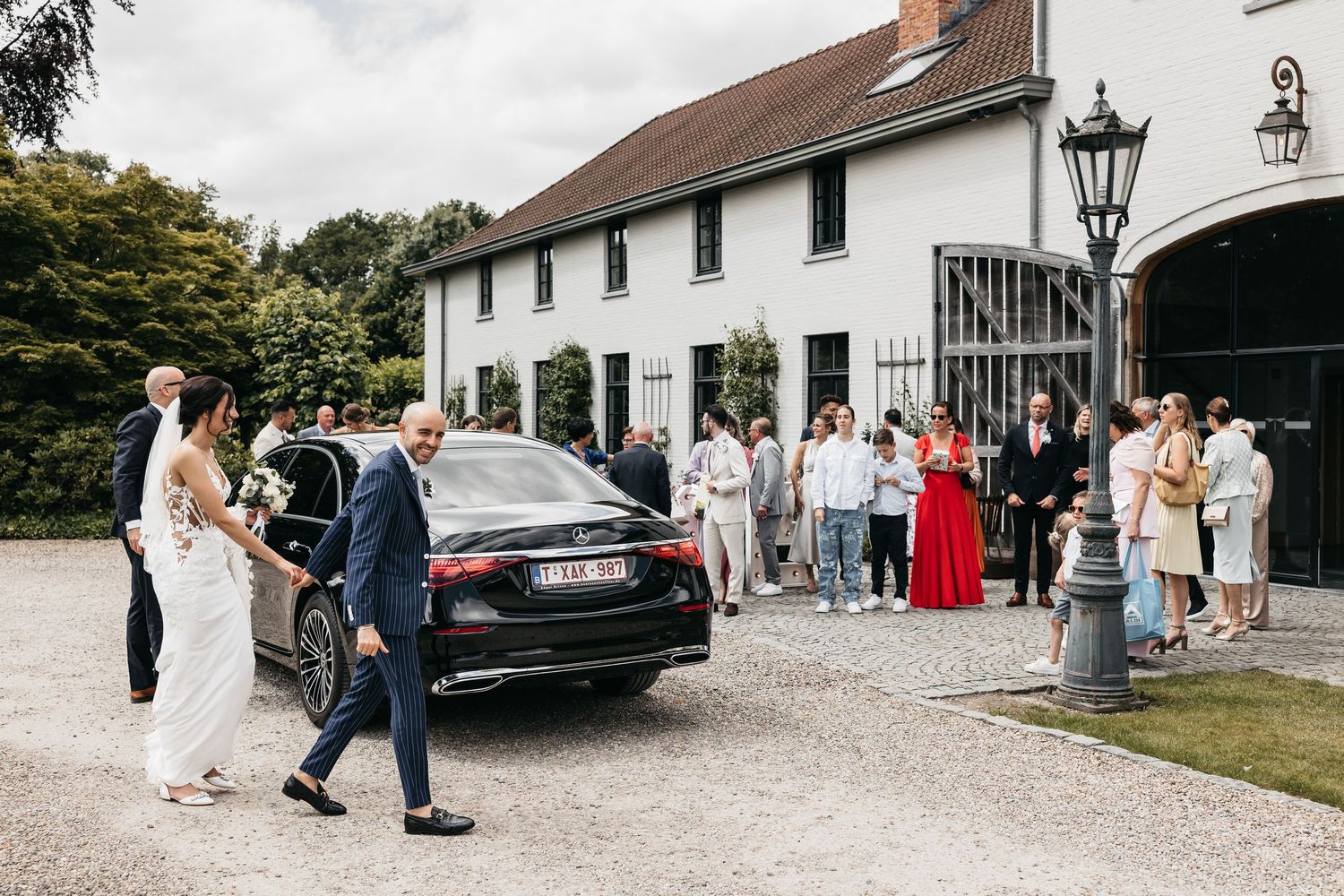 Bride and escort walk toward black wedding car as guests gather outside elegant white venue.