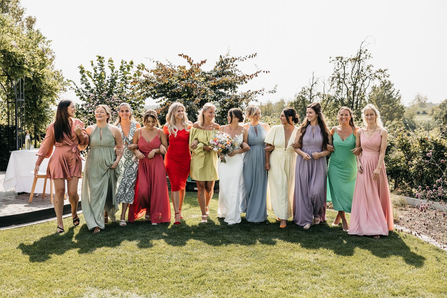Bride poses with ten bridesmaids wearing colorful mismatched dresses on a sunny outdoor garden wedding venue.