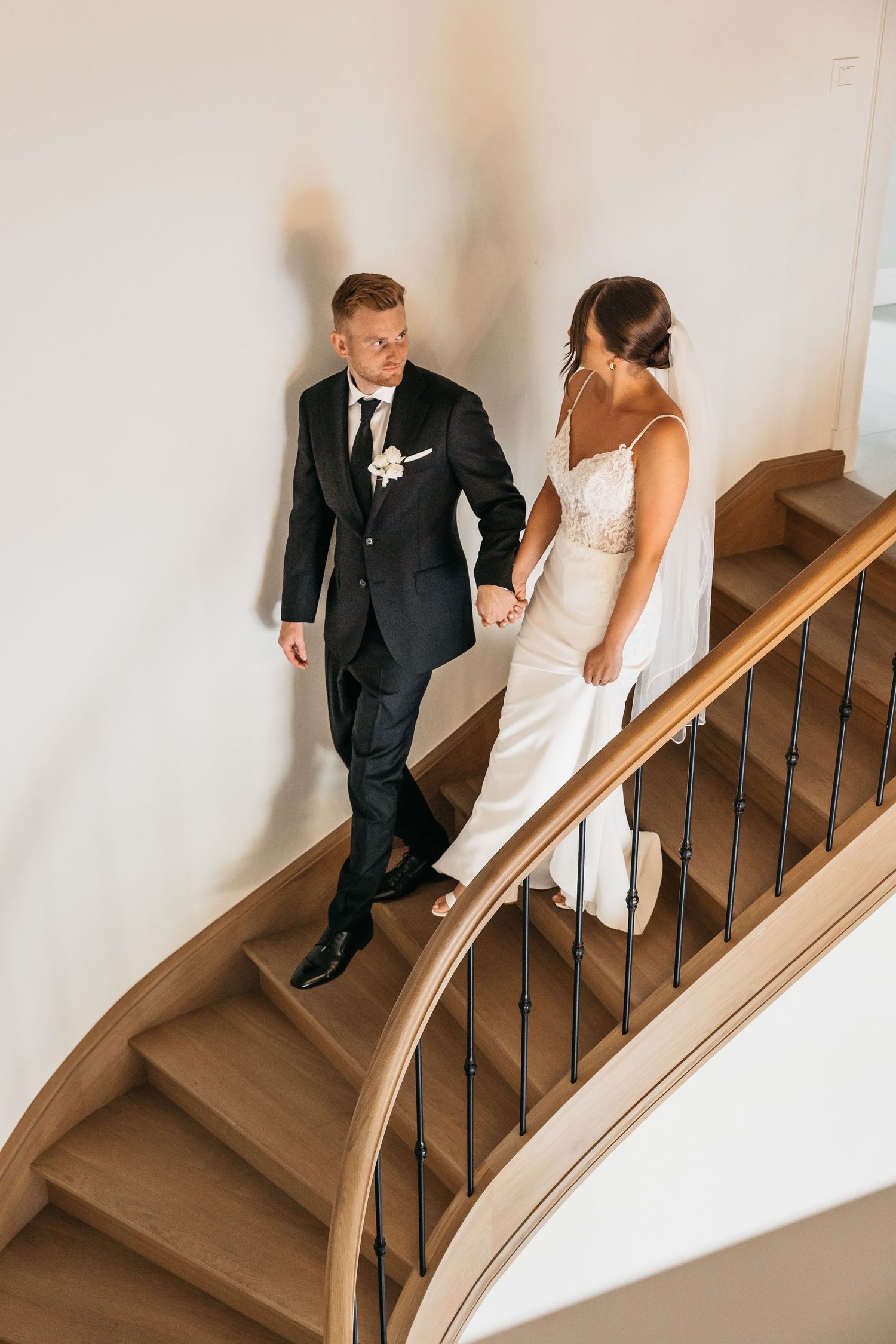Bride and groom holding hands while walking down curved wooden staircase in wedding attire.