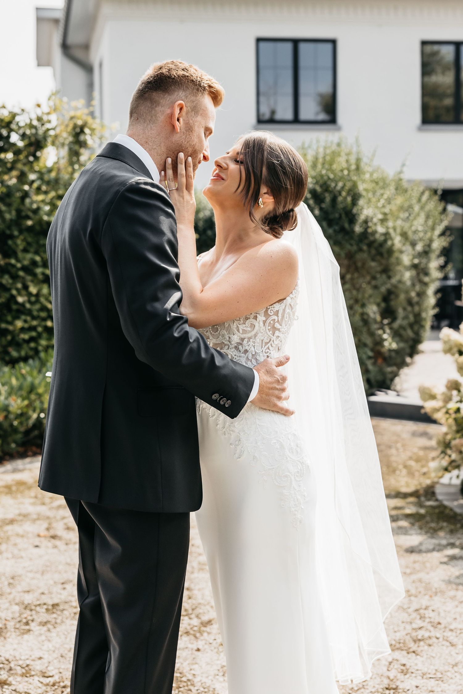 Bride and groom gaze lovingly at each other while embracing in sunny garden setting.