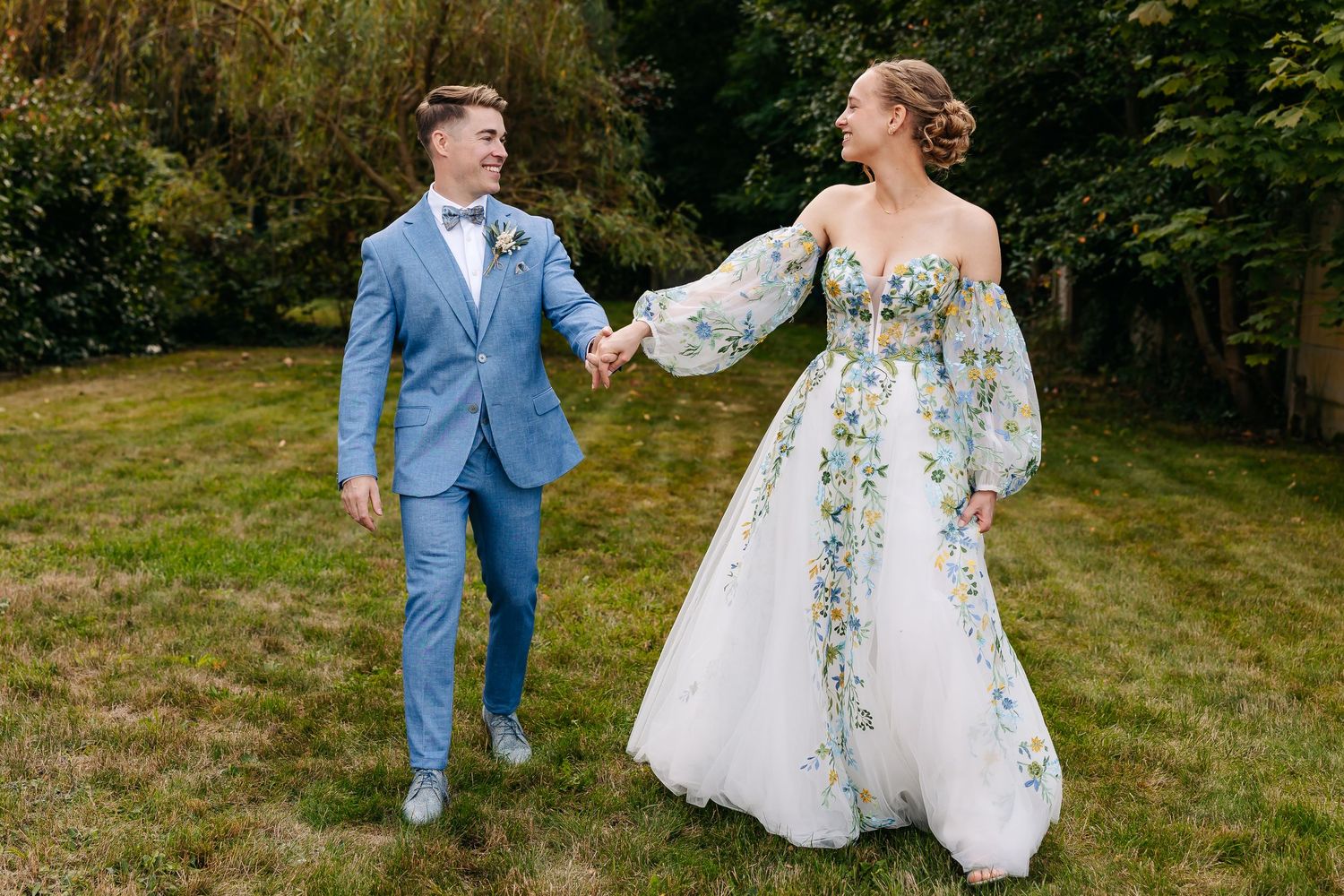 Happy newlyweds walk hand-in-hand on green lawn, bride in white floral gown and groom in light blue suit.