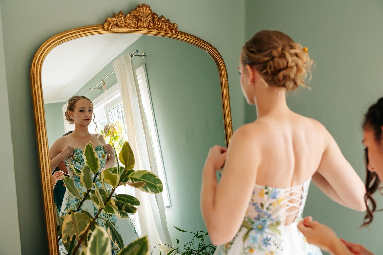 Smiling bride in floral strapless gown admiring her reflection in ornate gold-framed mirror.
