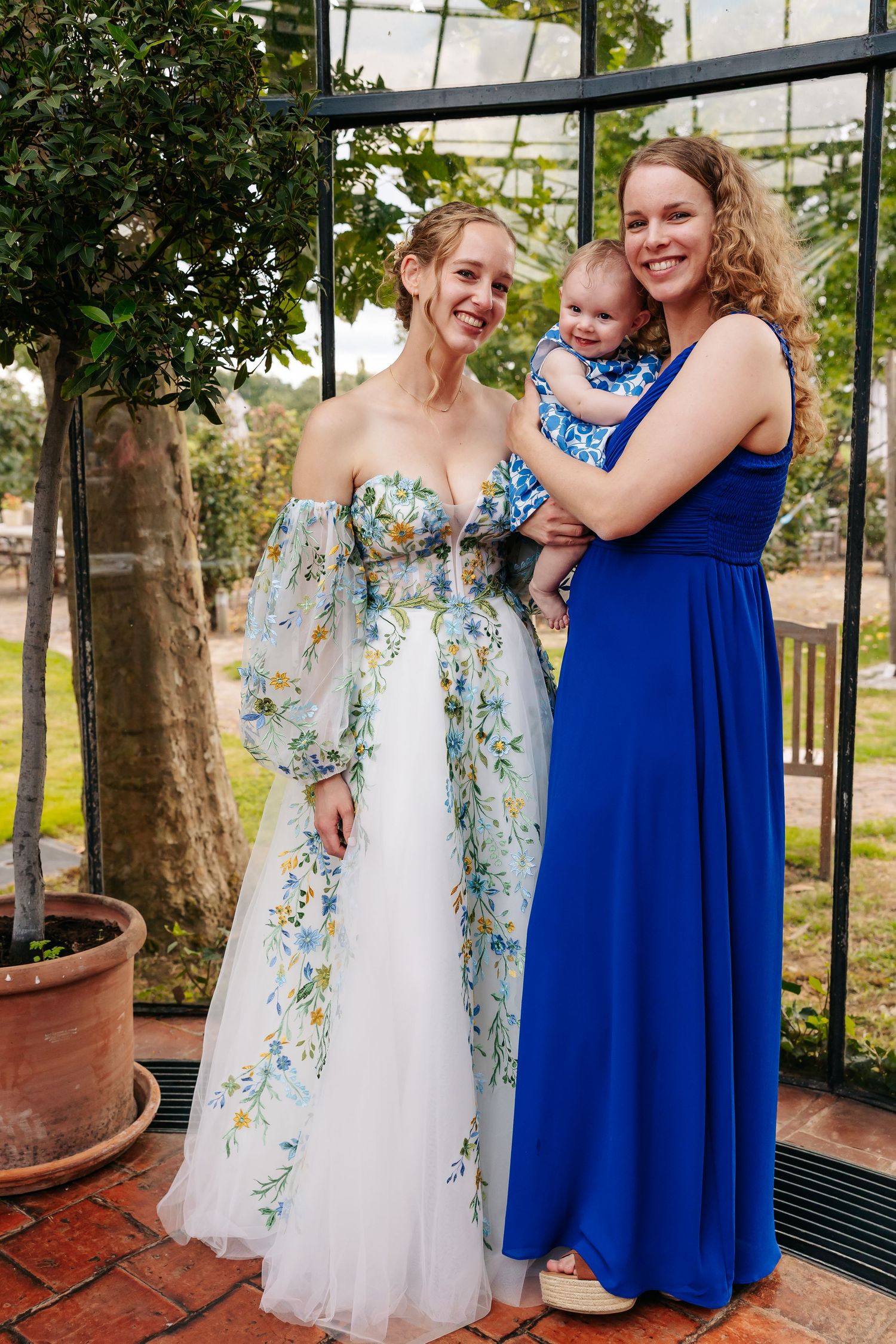 A bride in a floral gown poses with a woman in blue holding a baby in a greenhouse.