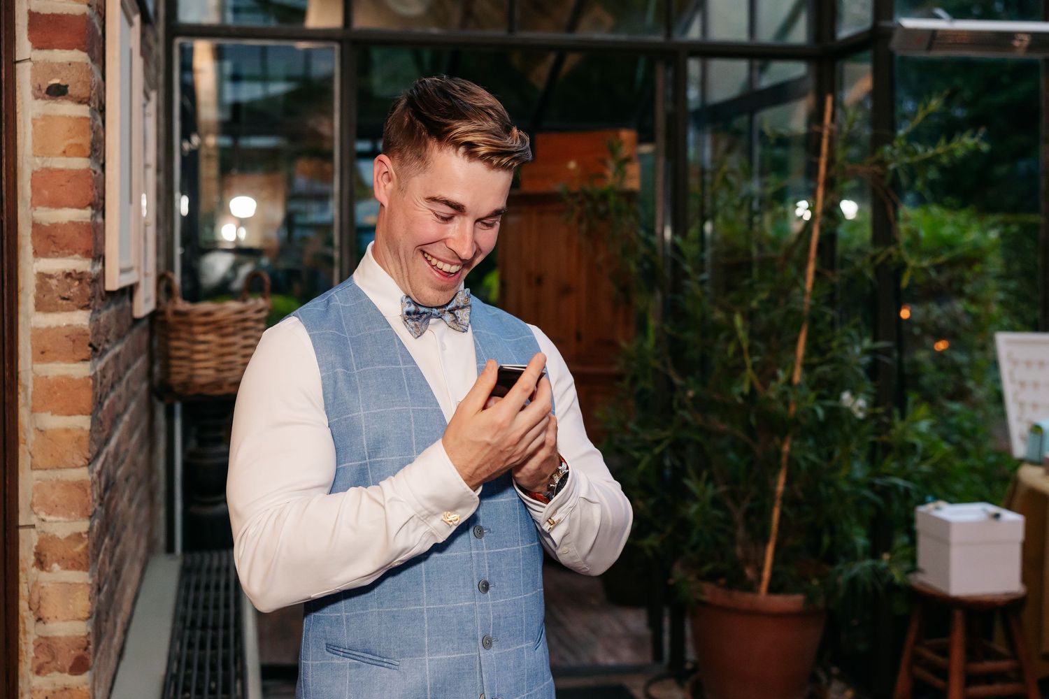 Smiling groom in blue vest and bow tie checking his phone at a rustic venue.