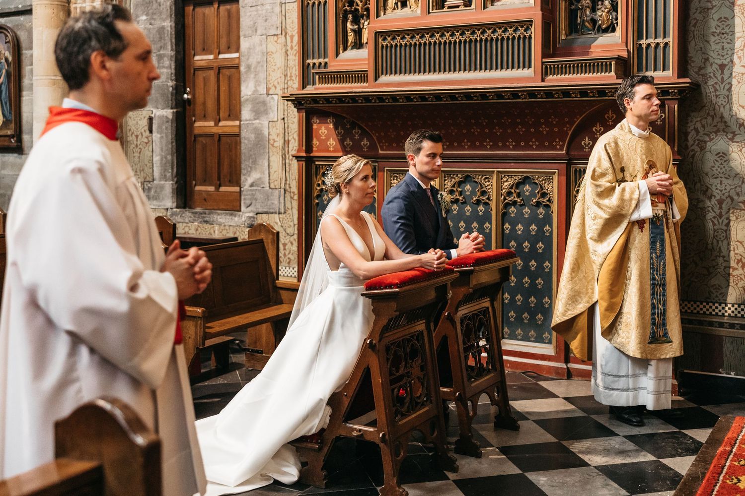 Bride and groom seated at ornate wooden altar during traditional Catholic church wedding ceremony.